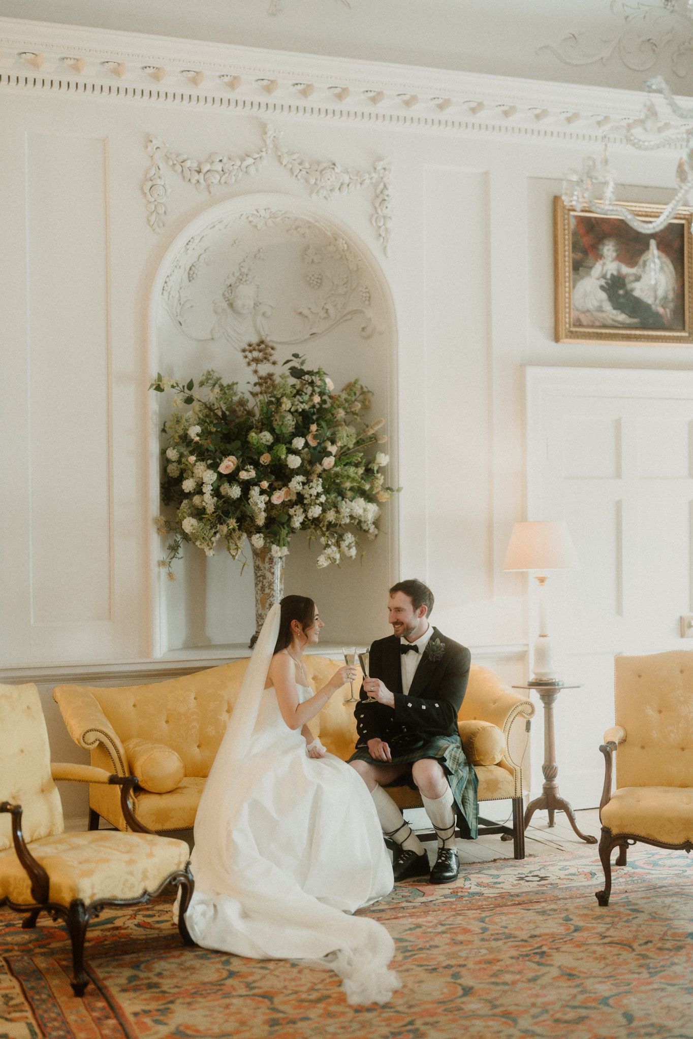 Bride and groom sitting on yellow sofa in elegant white room with floral arrangement, sharing champagne at their wedding.