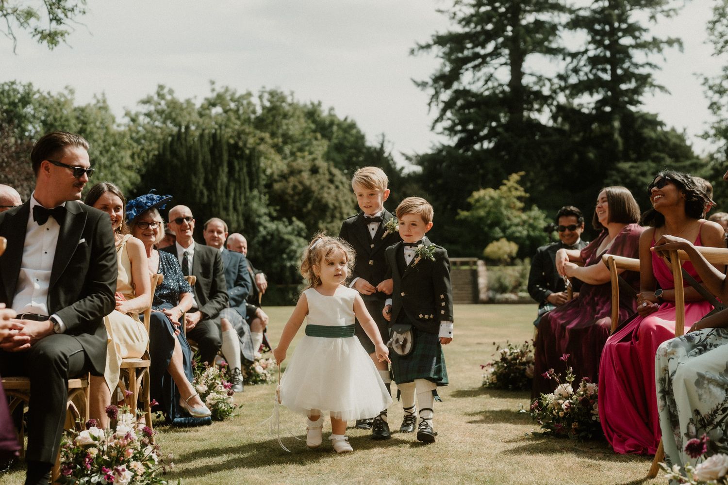 Flower girl in a white tulle dress and a young page boy in a kilt walk down the aisle as seated wedding guests watch outdoors.