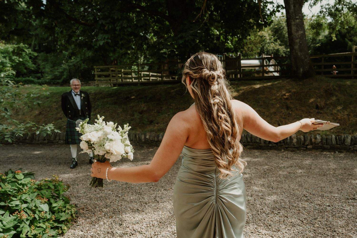 Bridesmaid in a sage green dress with a braided updo holds a white bouquet while an older man in a kilt watches from a gravel path.