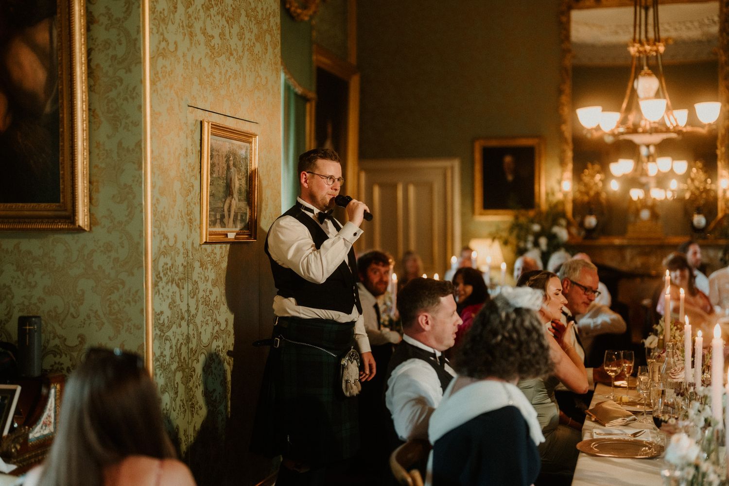 Man in a vest and kilt speaks into a microphone during a candlelit wedding dinner as seated guests listen in an elegant green-walled room.