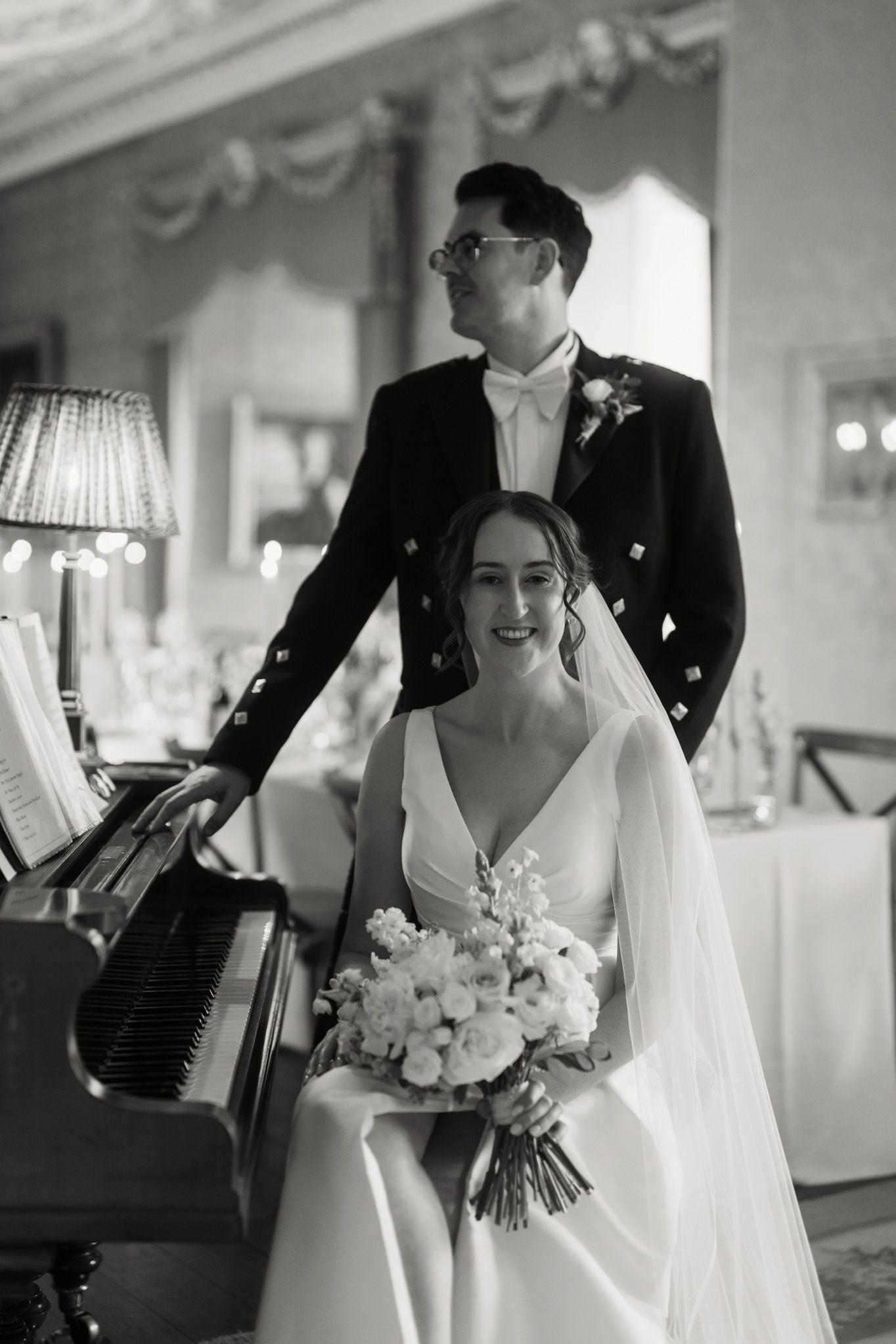 Bride in a white gown and veil holds a bouquet while seated at a piano, with a groom in a kilt jacket standing behind her.