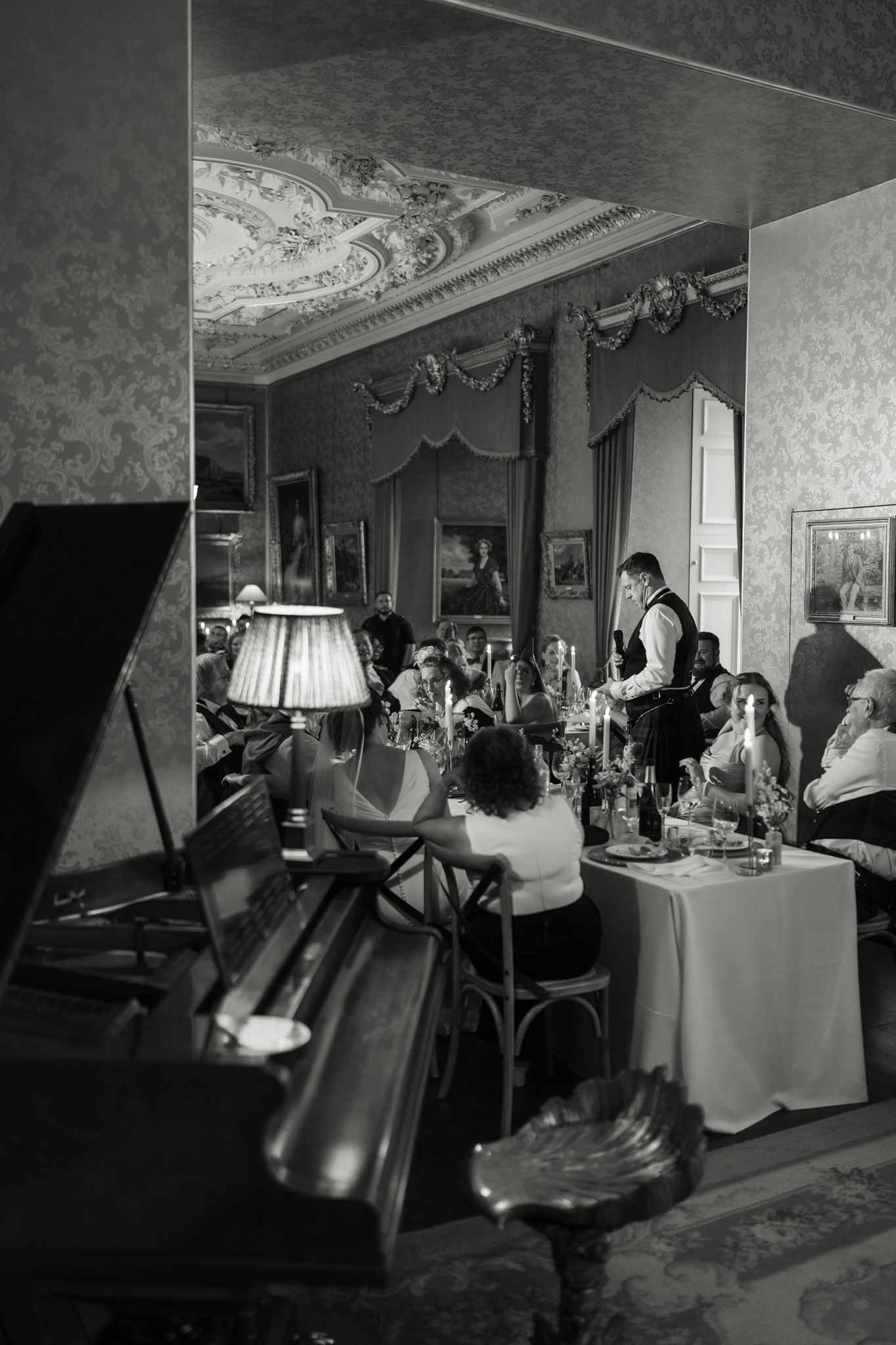Black and white photo of guests dining in an ornate room with a grand piano, lamp, draped walls and an elegant decorated ceiling.