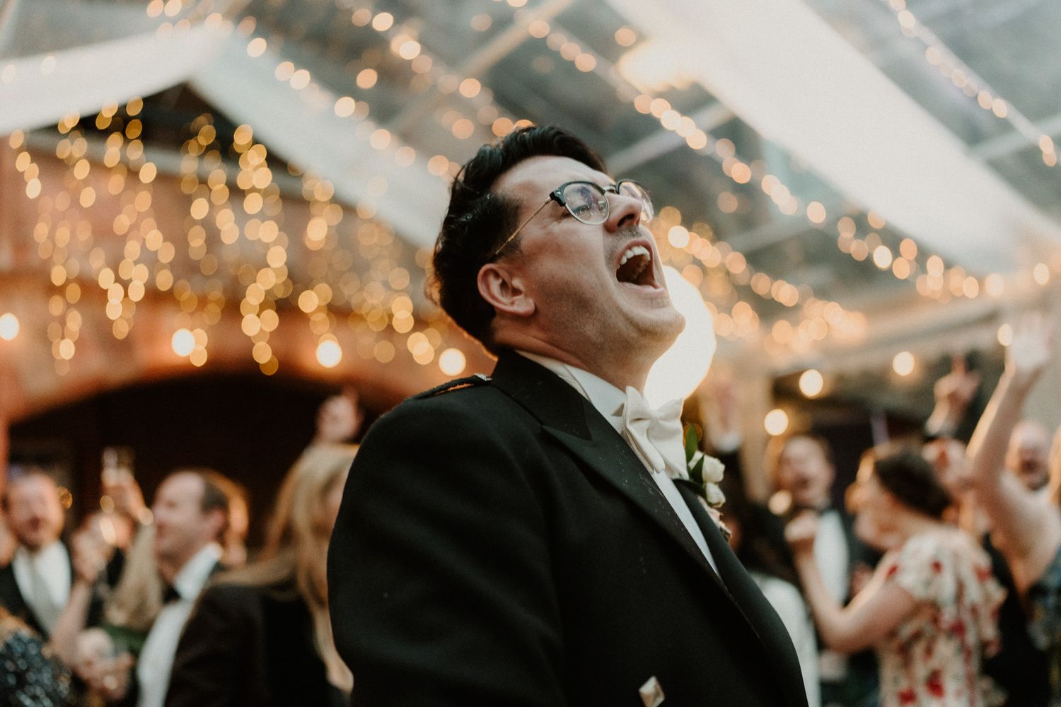 Man in glasses and dark formal jacket laughs joyfully with head tilted back at an evening wedding reception under glowing string lights.