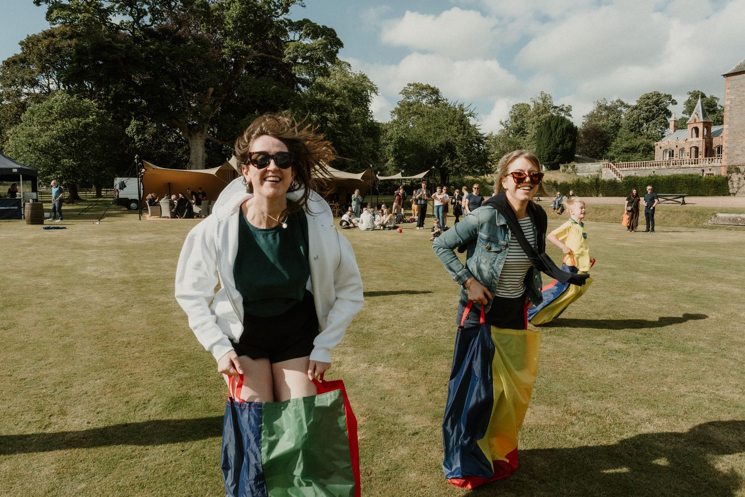 Two women in sunglasses laugh while standing in colorful sacks during a sack race on a sunny lawn at an outdoor summer festival.