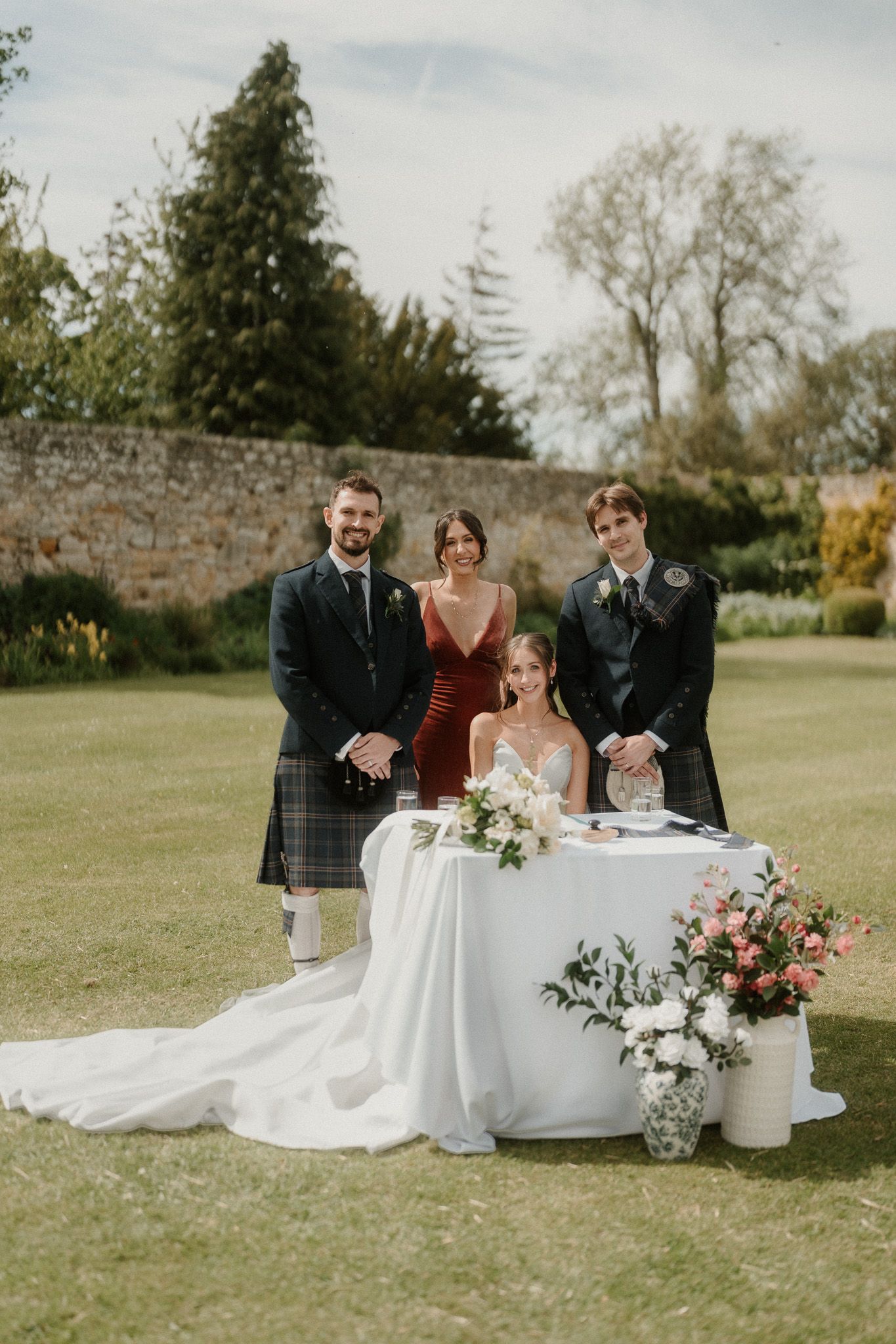 Bride in white gown seated at outdoor wedding table with groom in tartan kilt and guests in walled garden.