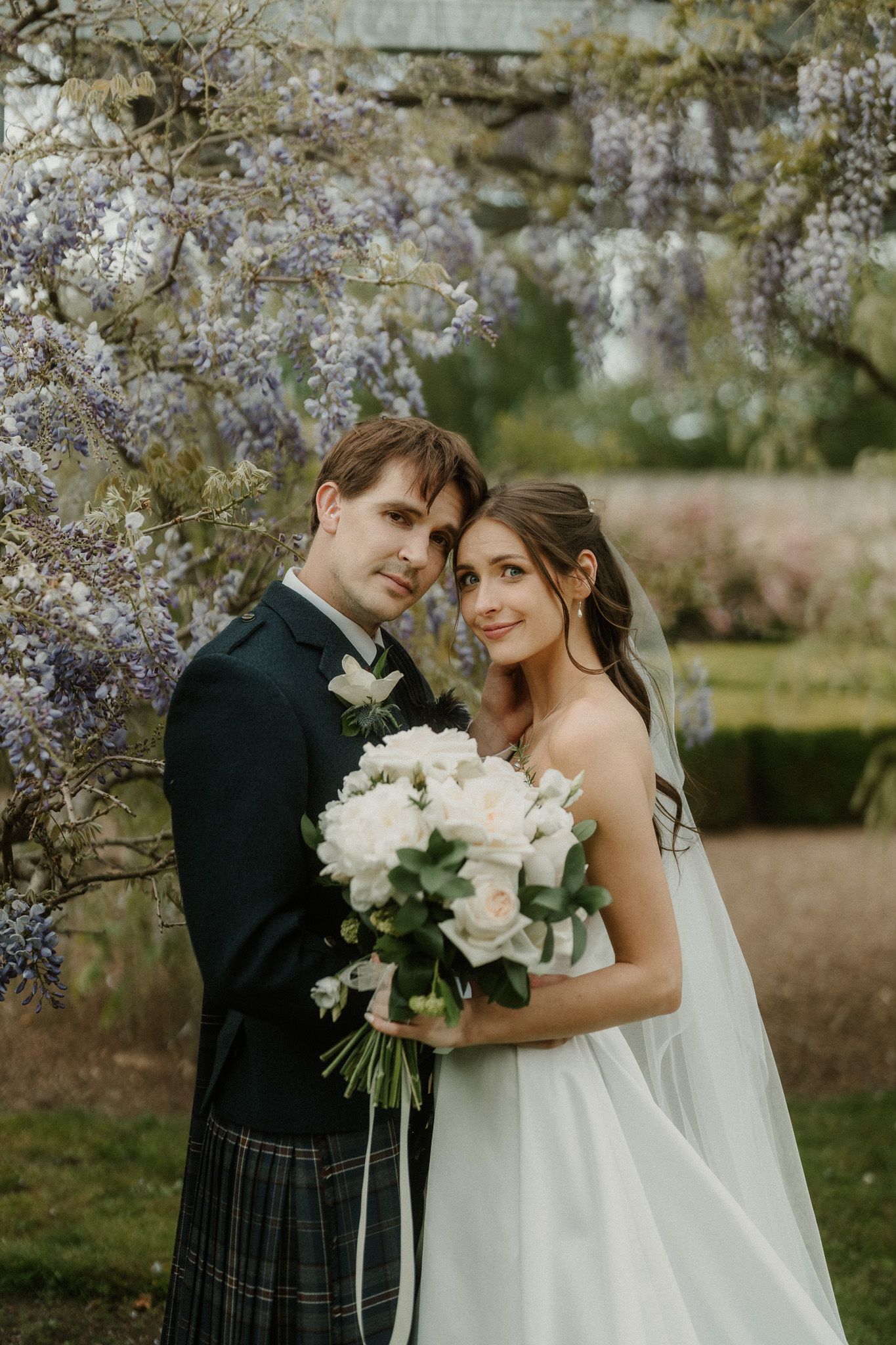 Bride and groom pose under blooming wisteria, bride holding white bouquet, groom in tartan kilt, garden wedding portrait.