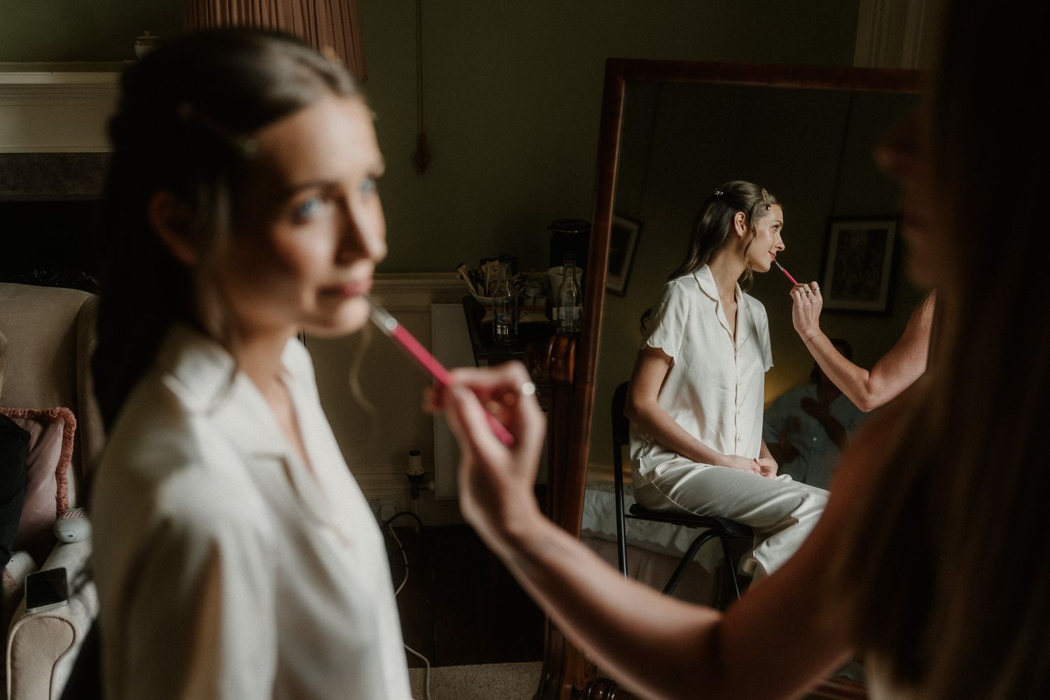 Bride getting makeup applied by artist, with mirror reflection visible in elegant bridal preparation room.