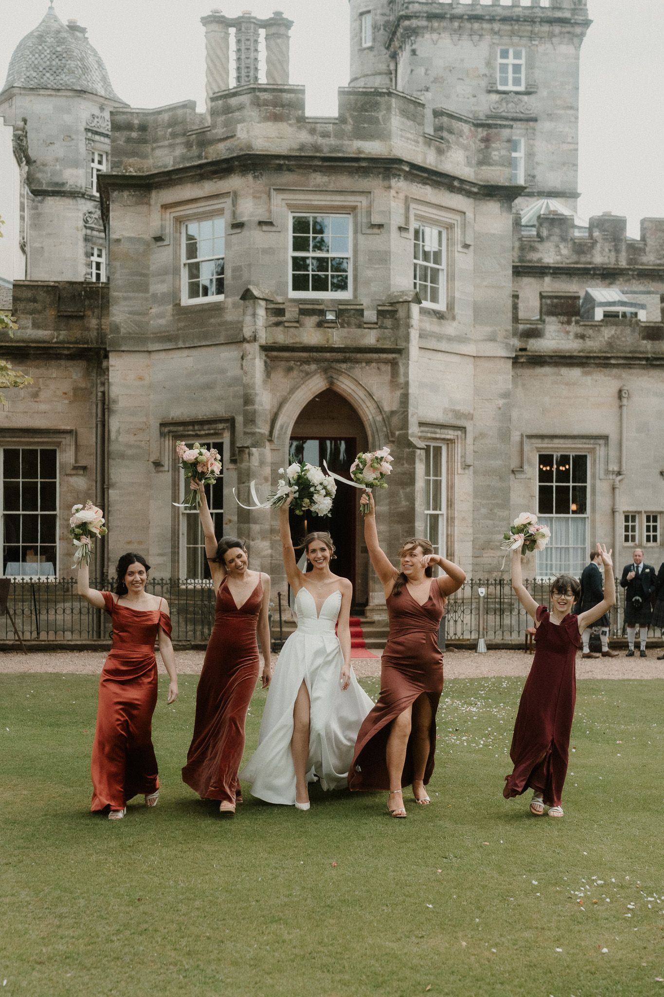 Bride in white gown celebrates with bridesmaids in burgundy dresses outside a grand stone castle venue.