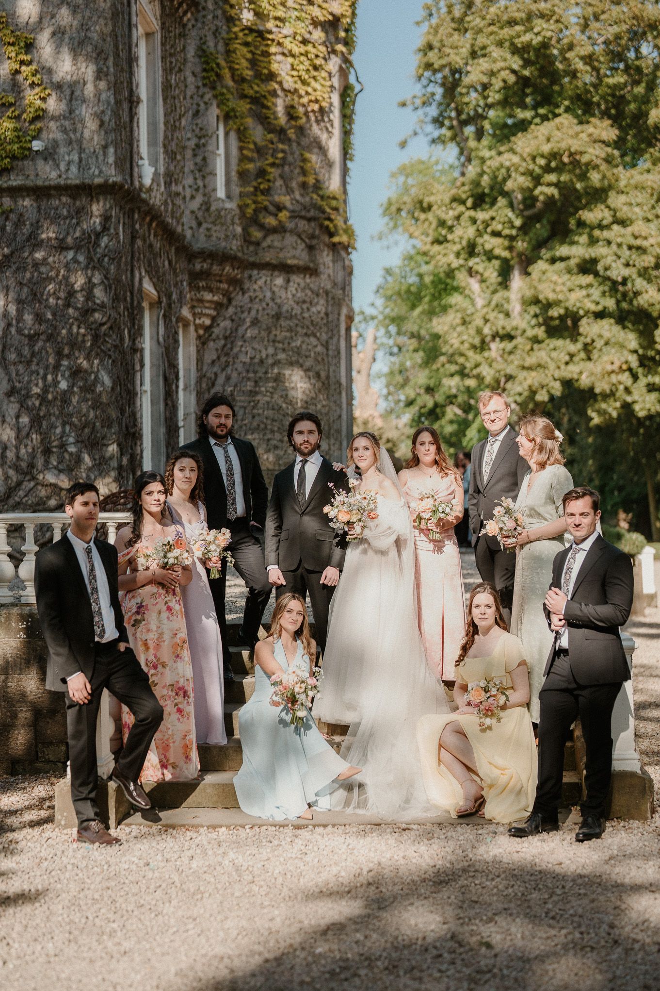Wedding party poses on stone steps outside ivy-covered castle, bride in white gown with bridesmaids holding floral bouquets.