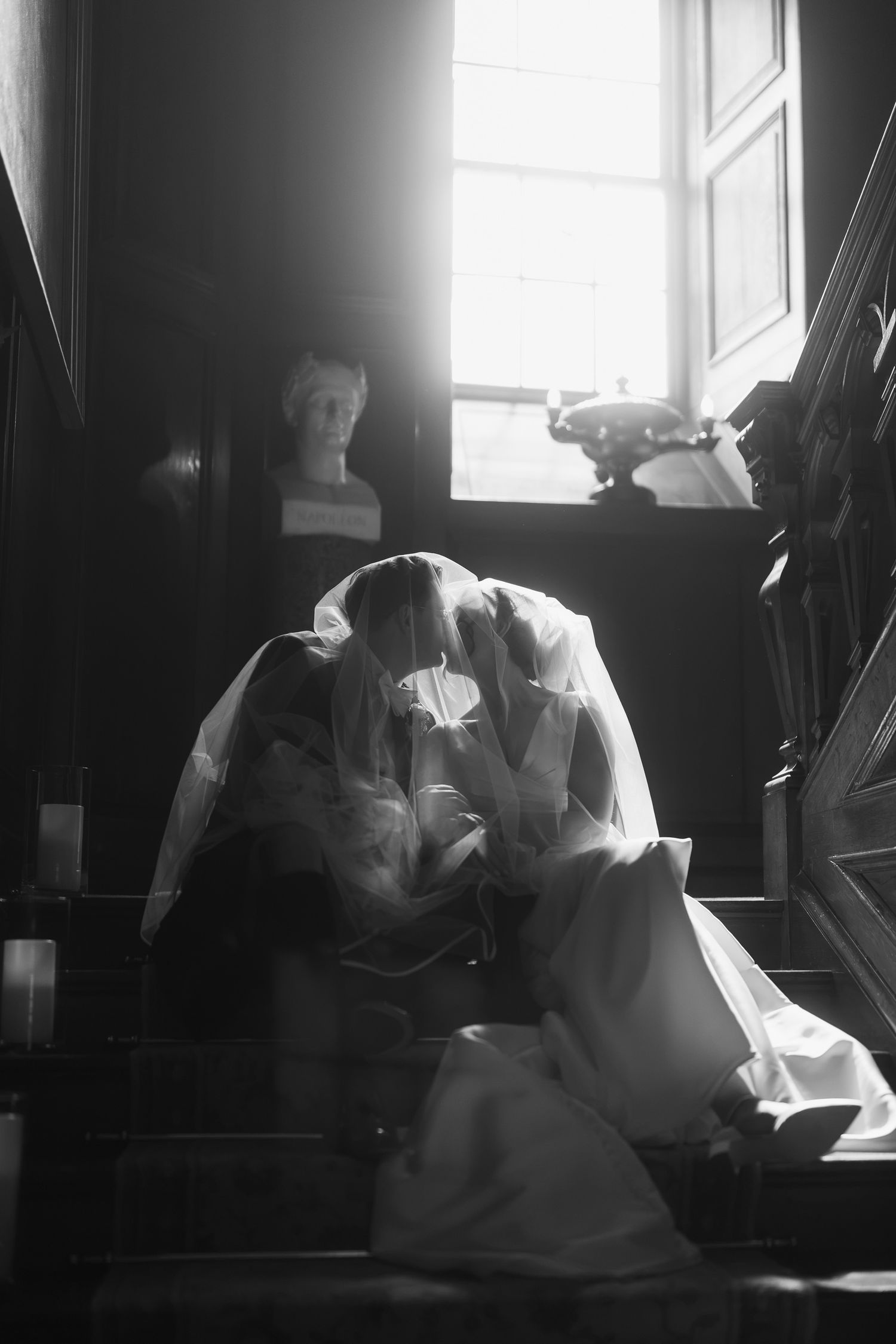 Two brides share an intimate moment on wooden stairs in a black and white wedding photo with soft window light.