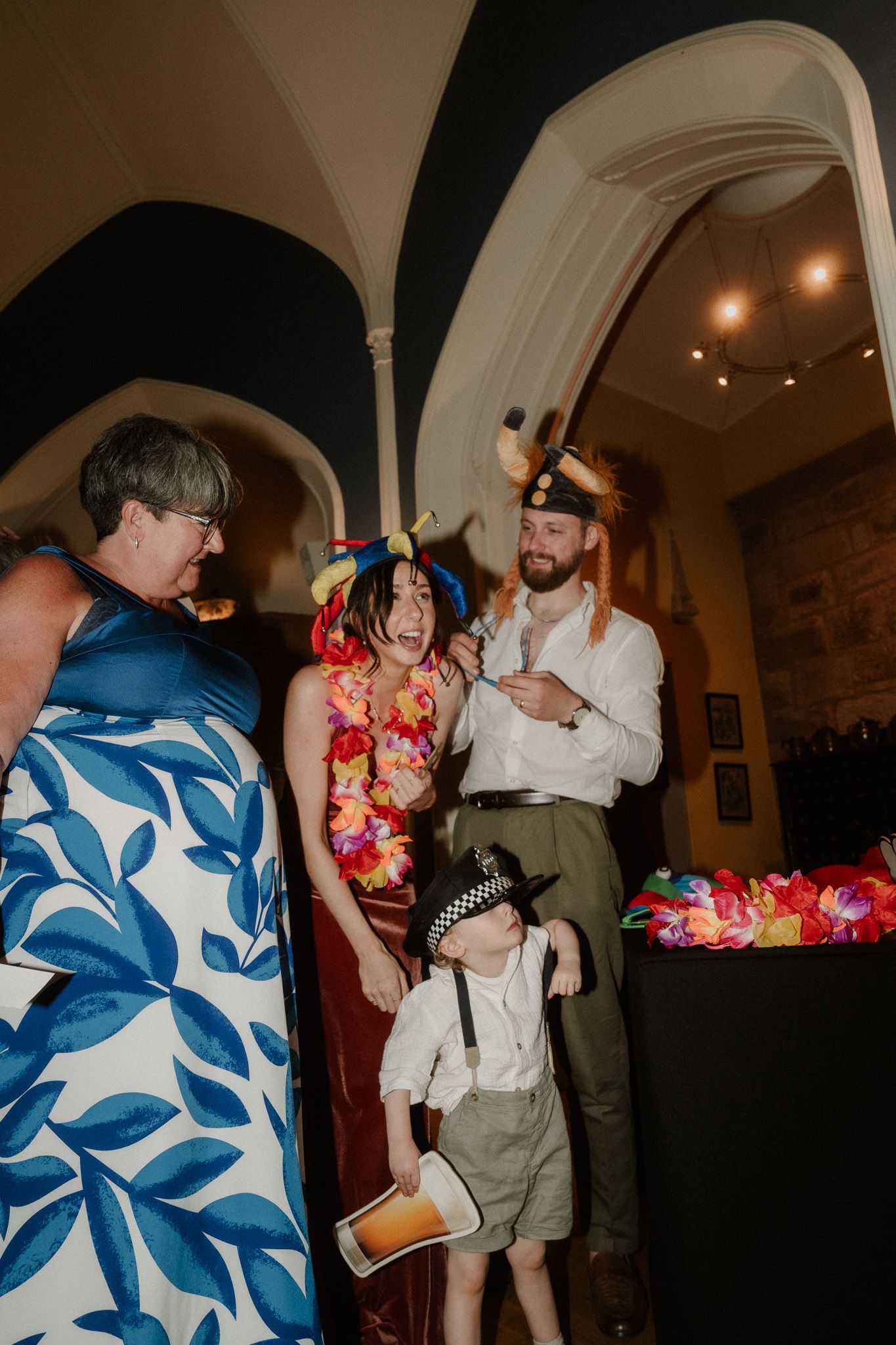Family in festive costumes laughing together at a wedding reception in an elegant arched venue.