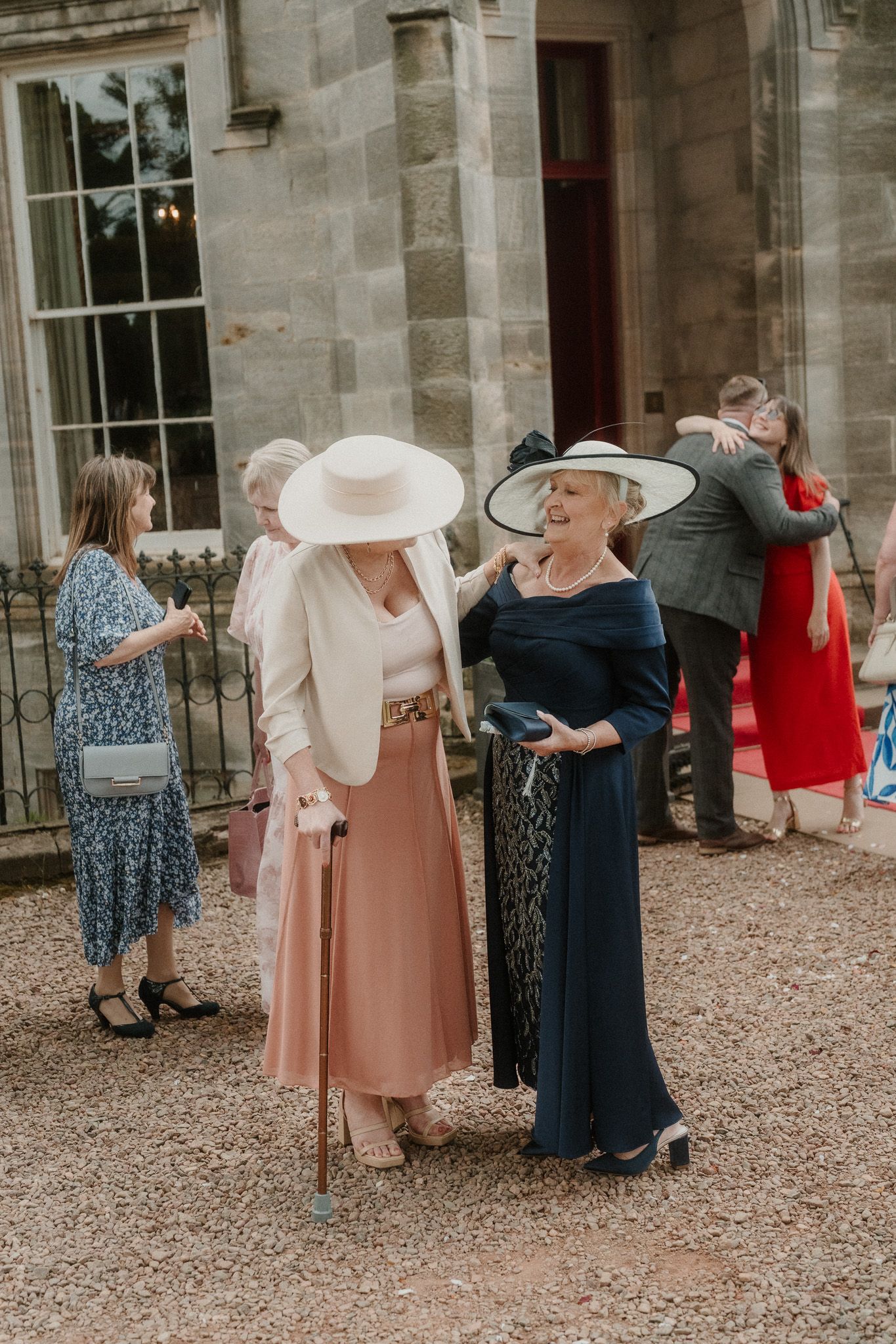 Two elegantly dressed women in wide-brim hats chat at an outdoor wedding reception near a stone manor house.