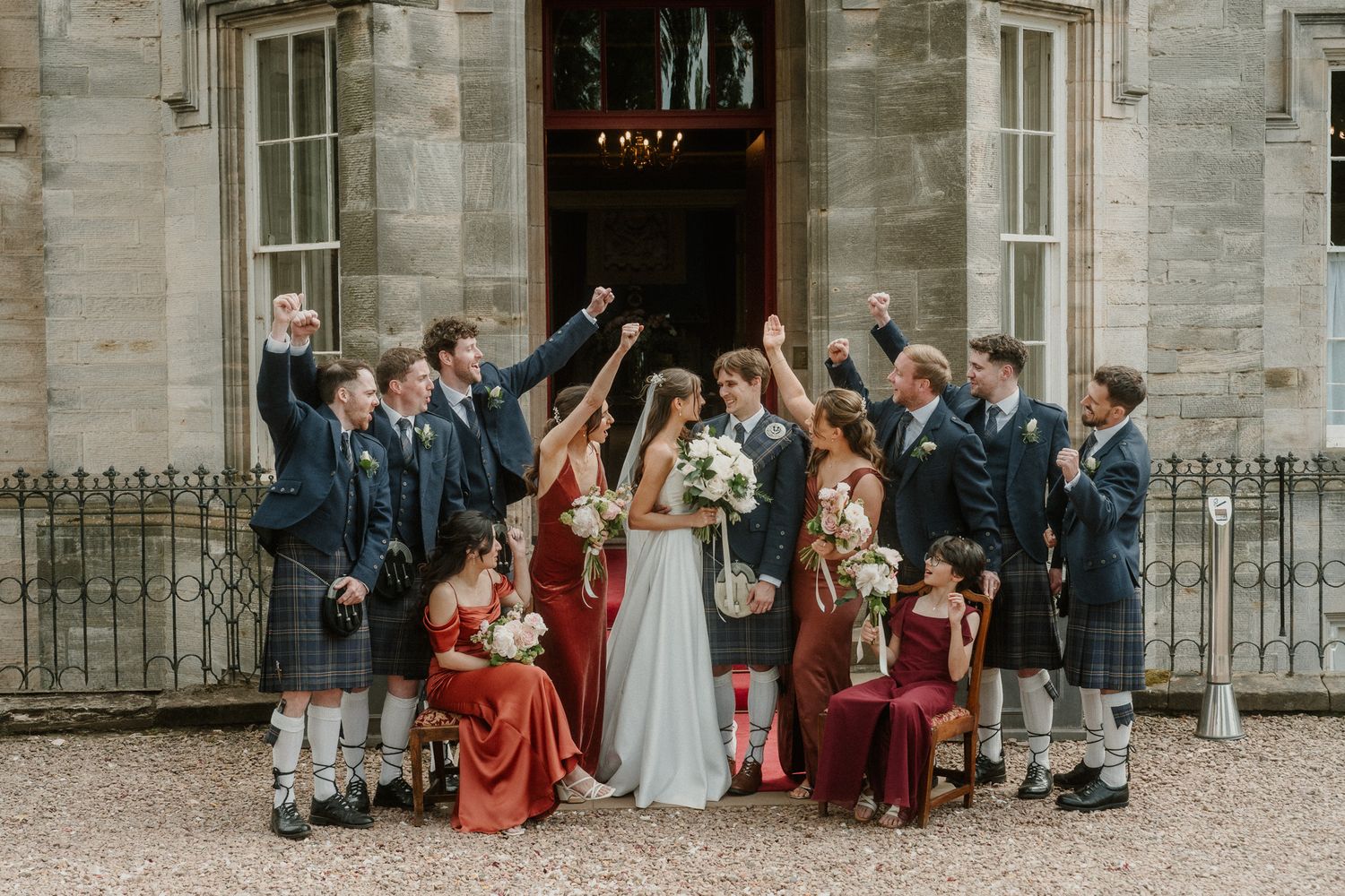 Wedding party celebrating outside a grand stone building, bridesmaids in red dresses and groomsmen in kilts cheering joyfully.