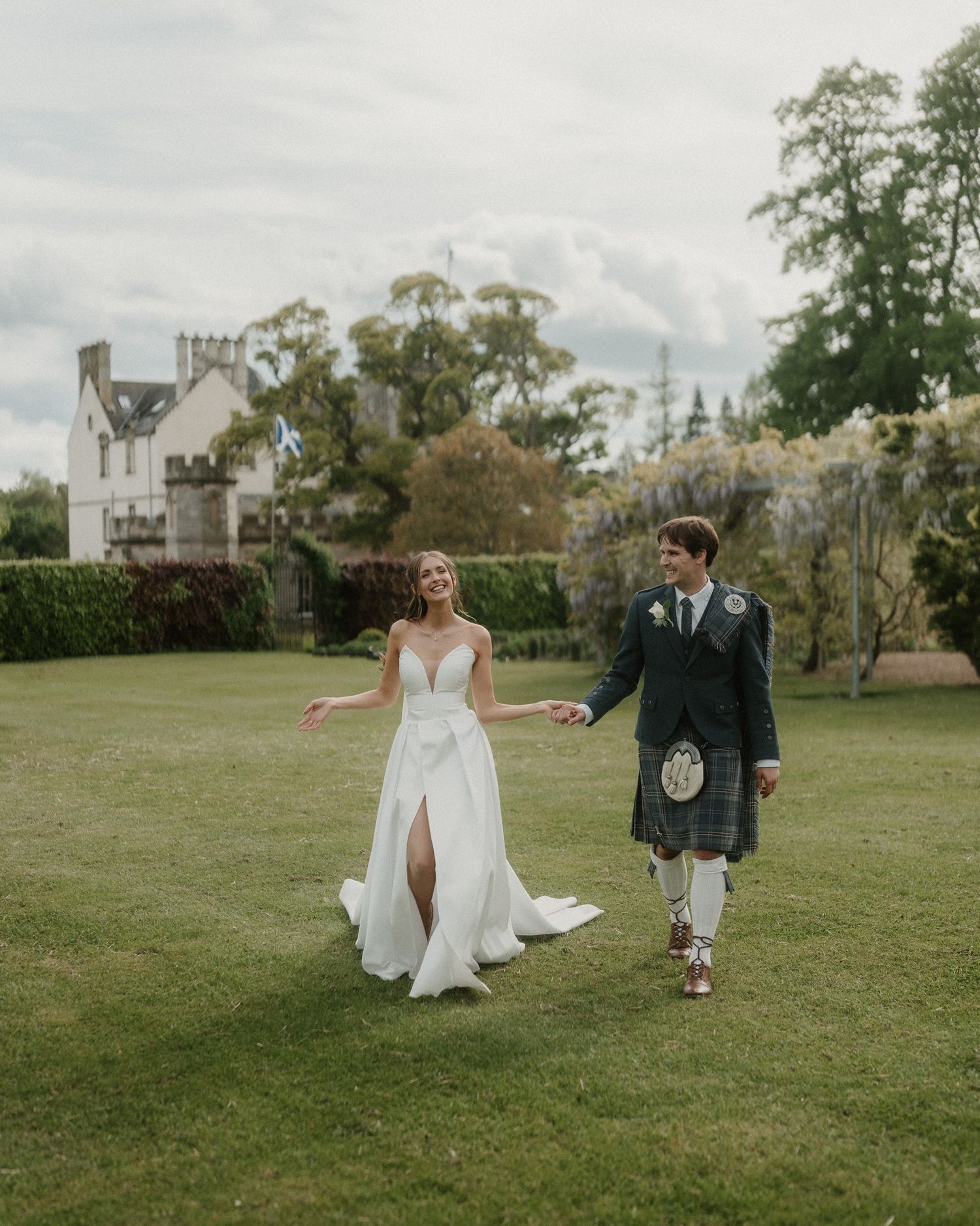 Bride in white gown and groom in Scottish kilt walk hand in hand on lush green lawn before a historic castle.