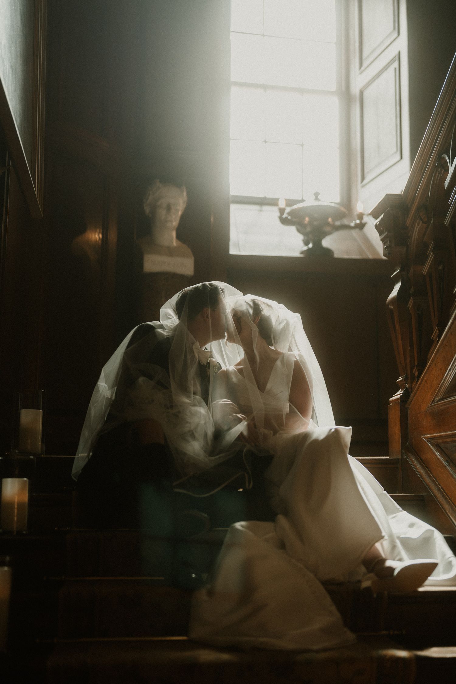 Bride and groom share a romantic kiss on wooden stairs, draped in a flowing veil, with soft window light and a marble bust.