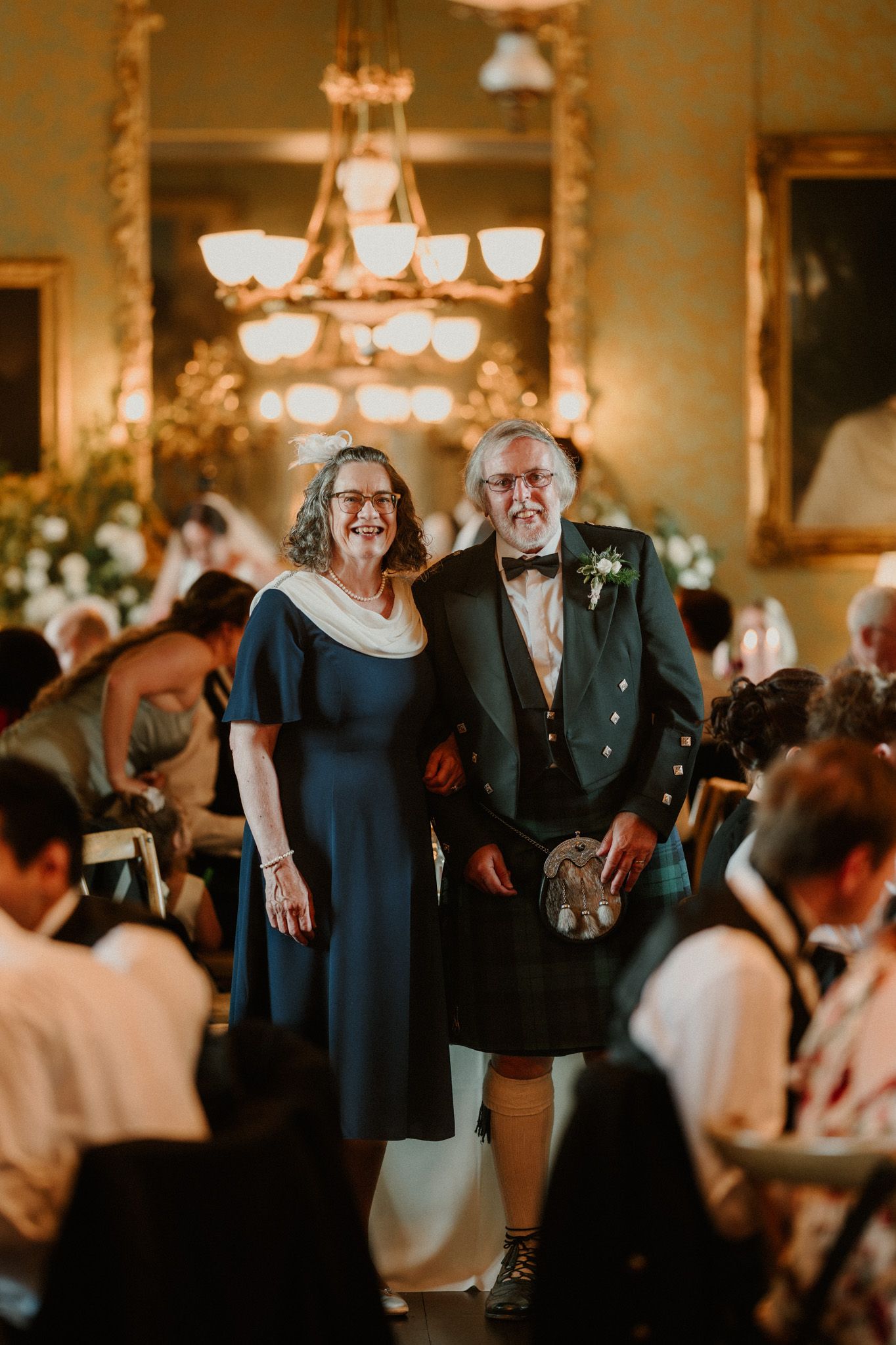 Smiling older couple arm in arm at a wedding reception, the man in a green tartan kilt and bow tie, the woman in a navy dress.