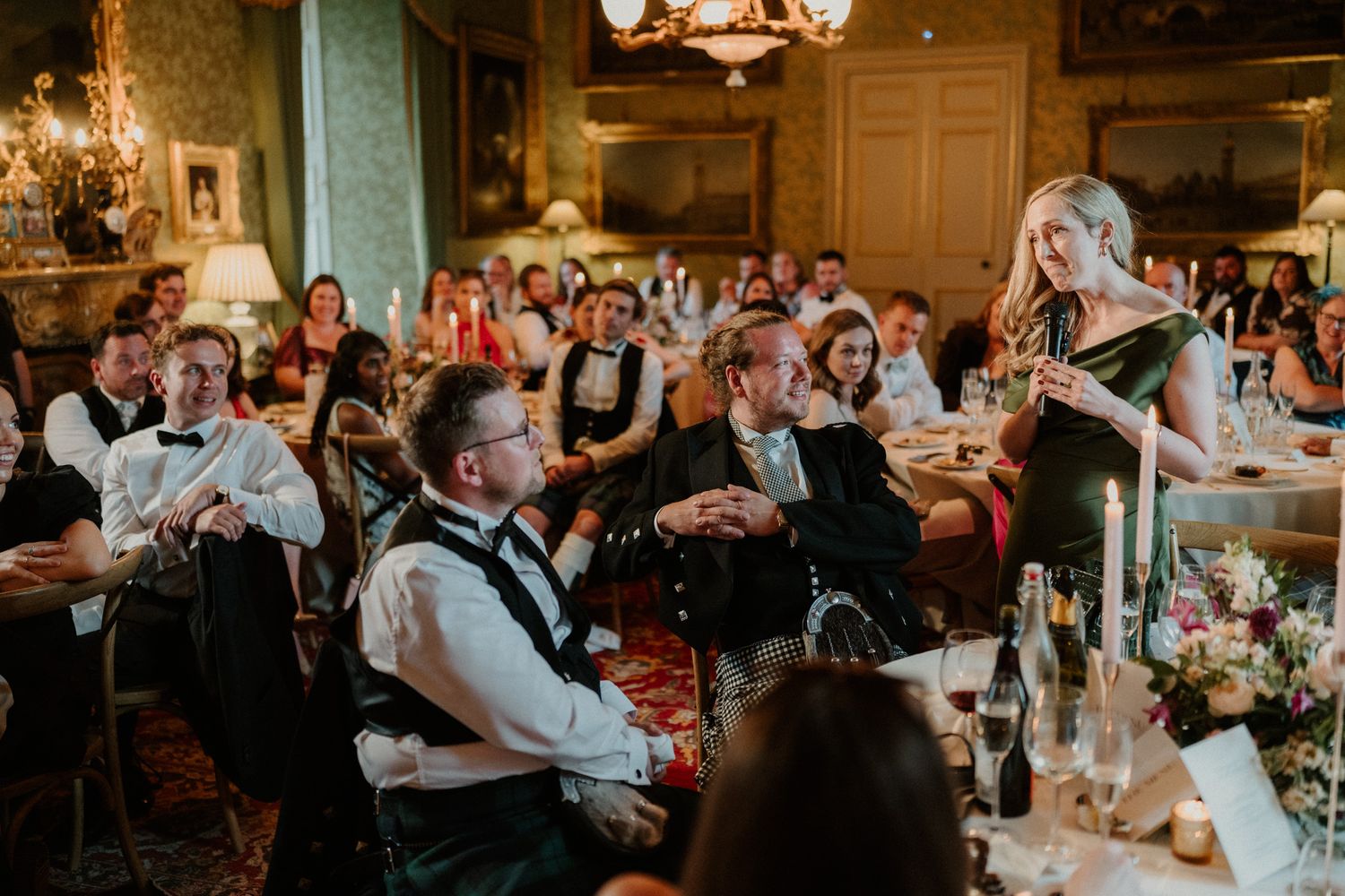 Woman in a dark dress gives a speech at an elegant wedding reception while seated guests in formal attire listen in an ornate room.