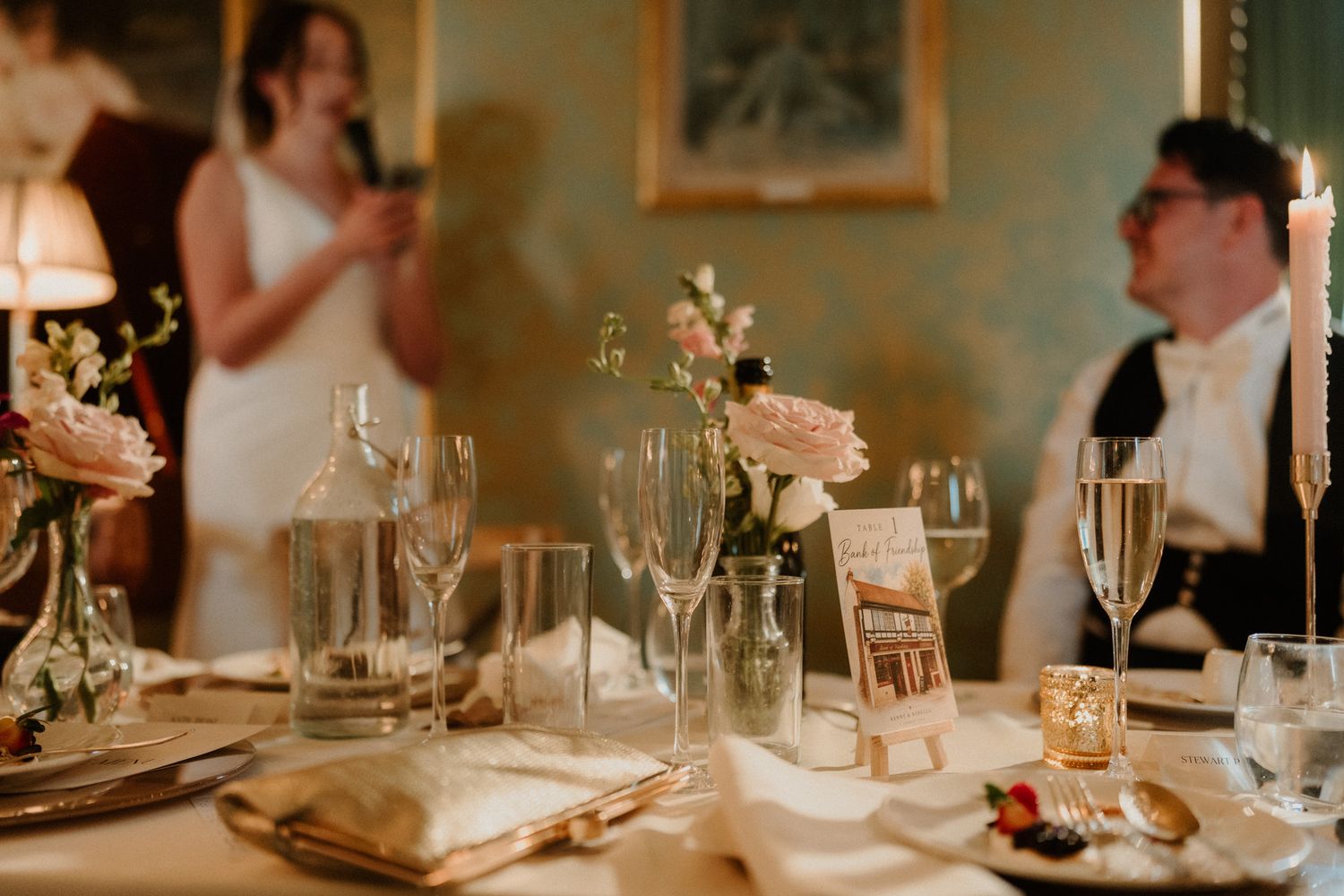 A wedding reception table with candles, pink roses, wine glasses and a place card, while a bride speaks behind a smiling guest.
