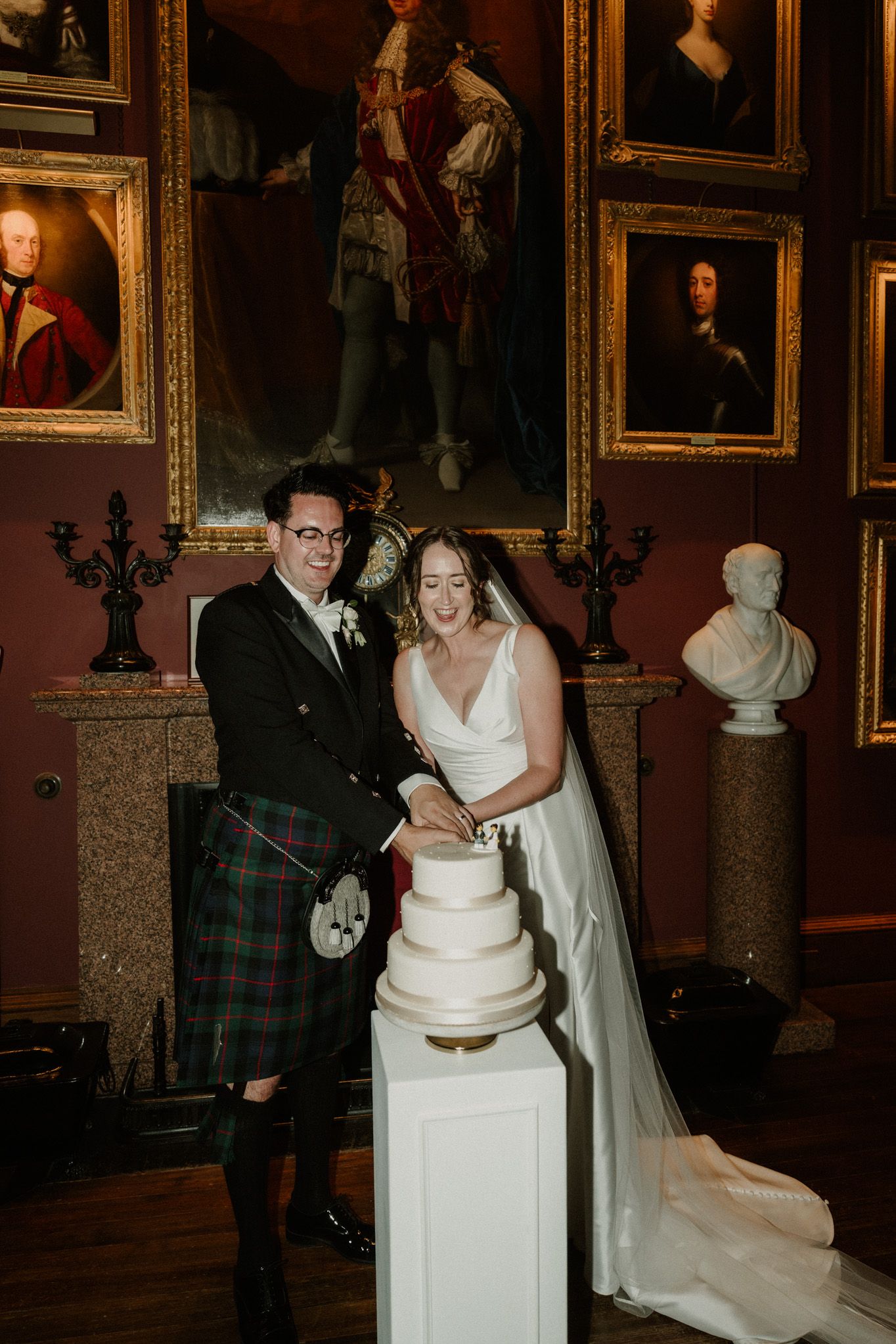 Bride in a white gown and veil and groom in a tartan kilt cut a three-tier white wedding cake in a gallery with framed portraits.