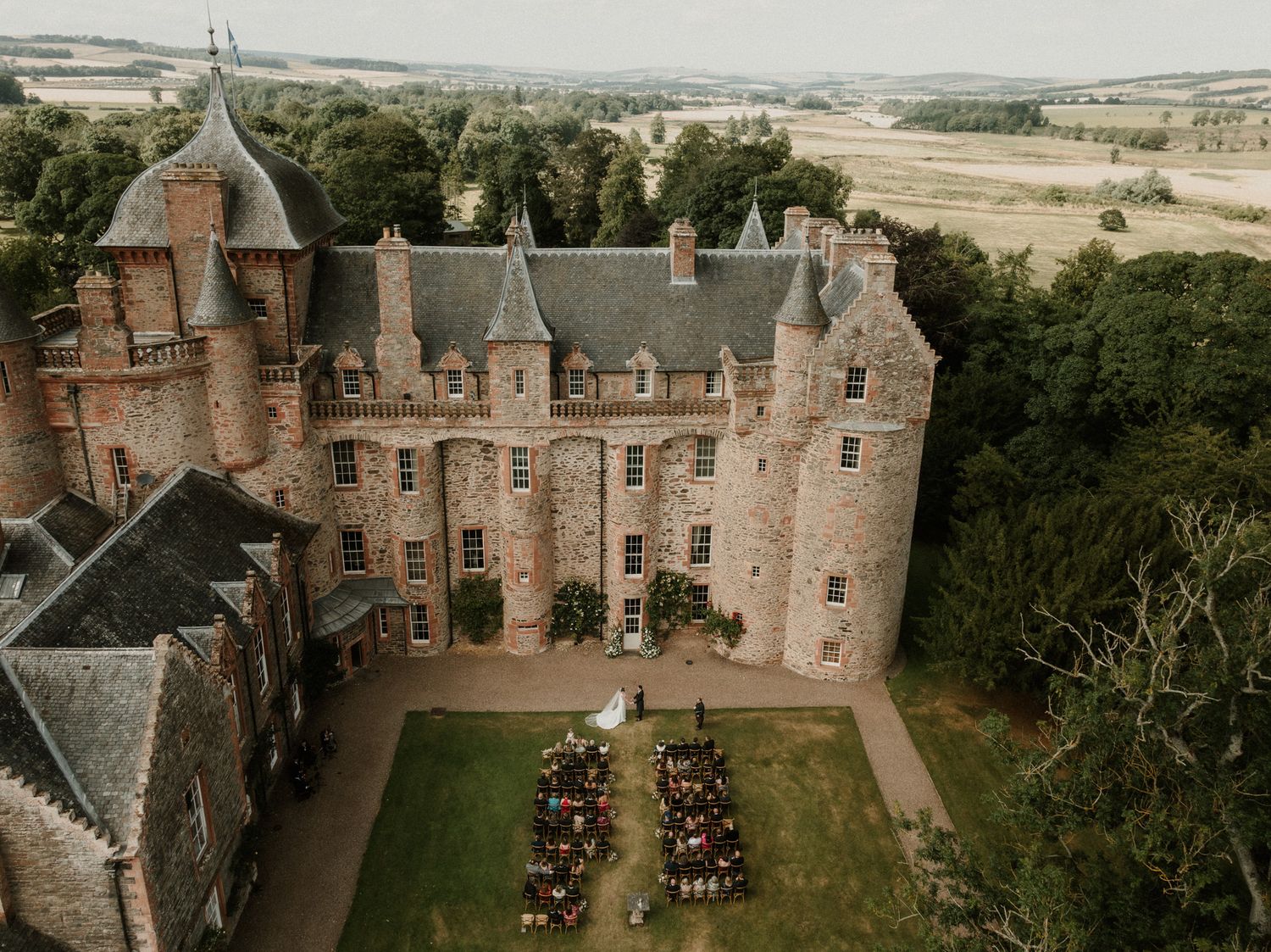 Aerial view of a historic stone castle with turrets and a courtyard lawn set up for an outdoor wedding ceremony with floral arrangements.