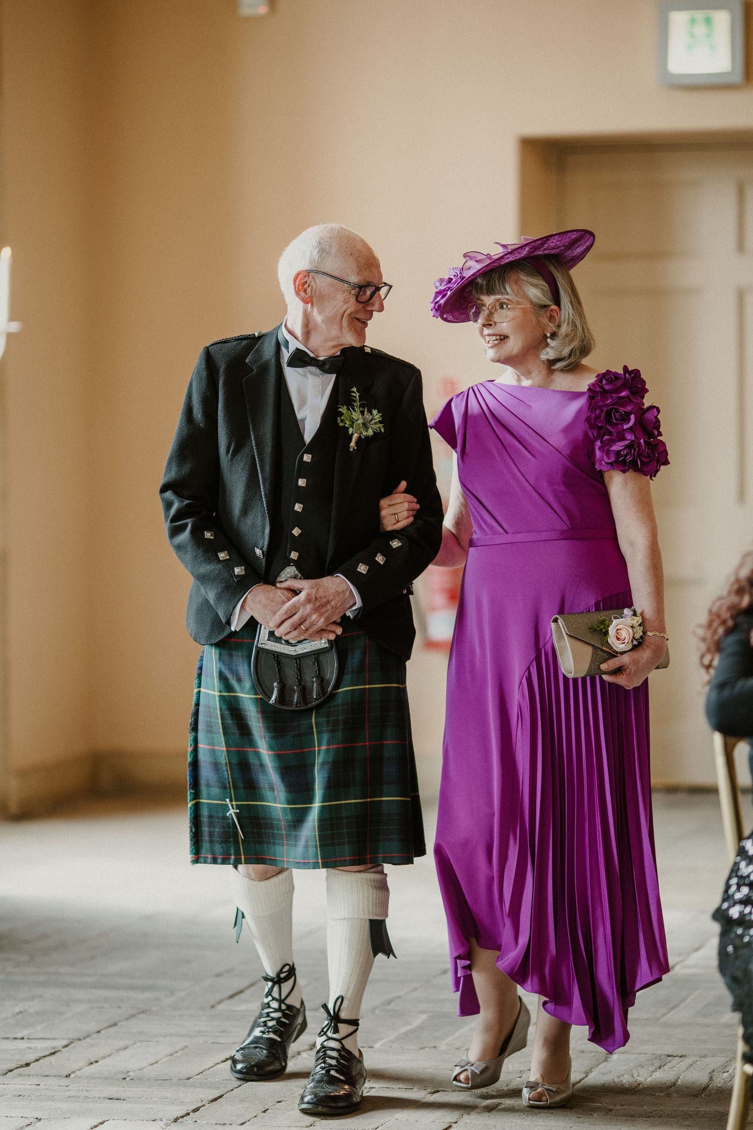 Wedding guests in Scottish kilts and purple formal attire gathered in an indoor venue.