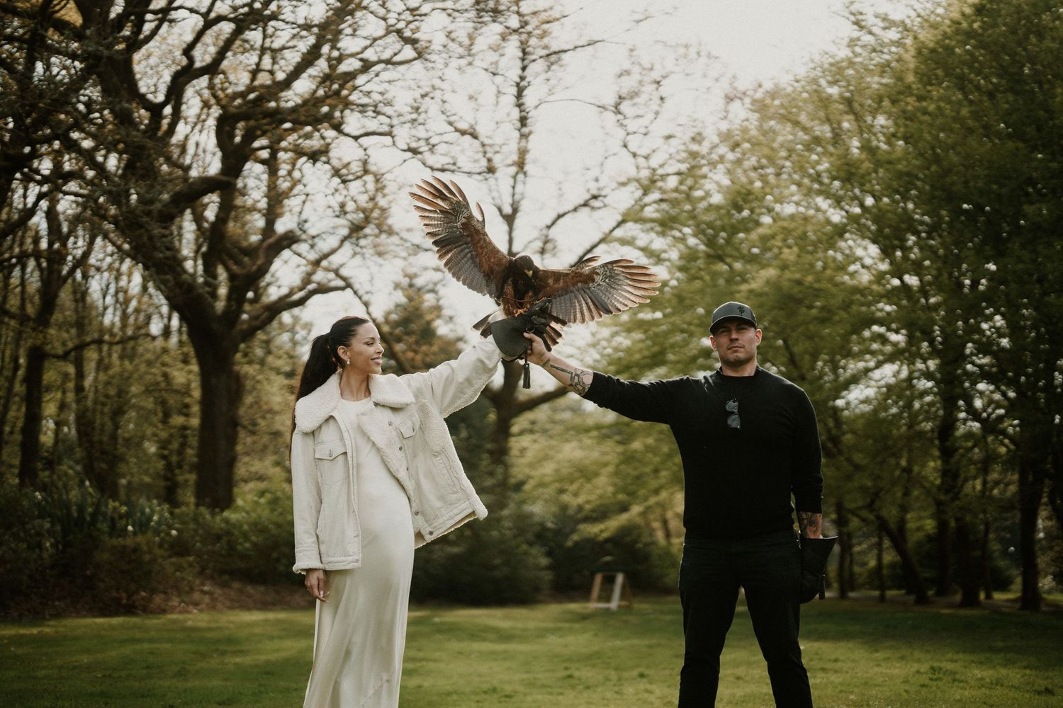A couple in formal attire releases an owl in flight between them in a wooded setting.