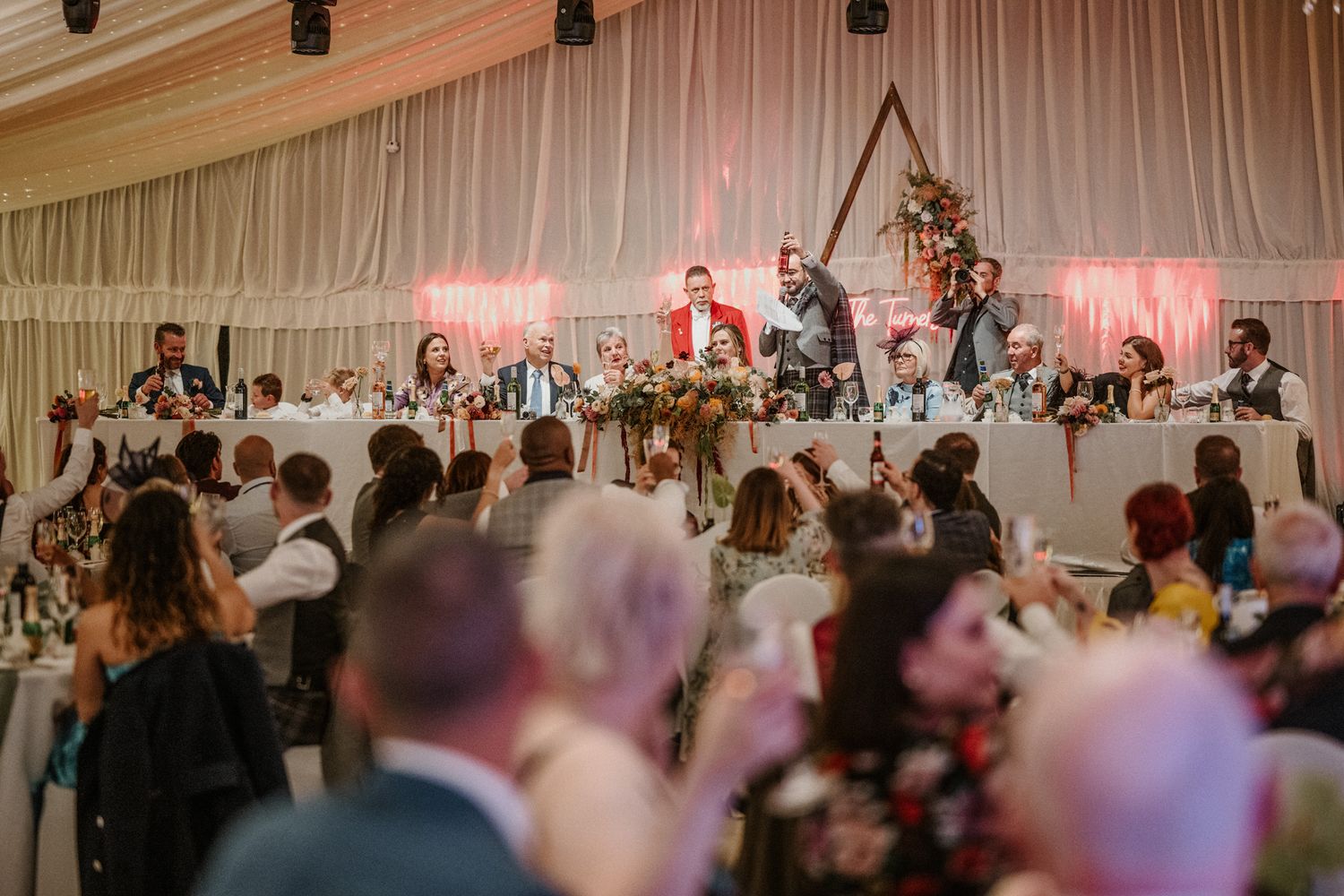 Crowded wedding reception with guests mingling under draped ceiling and pink accent lighting.