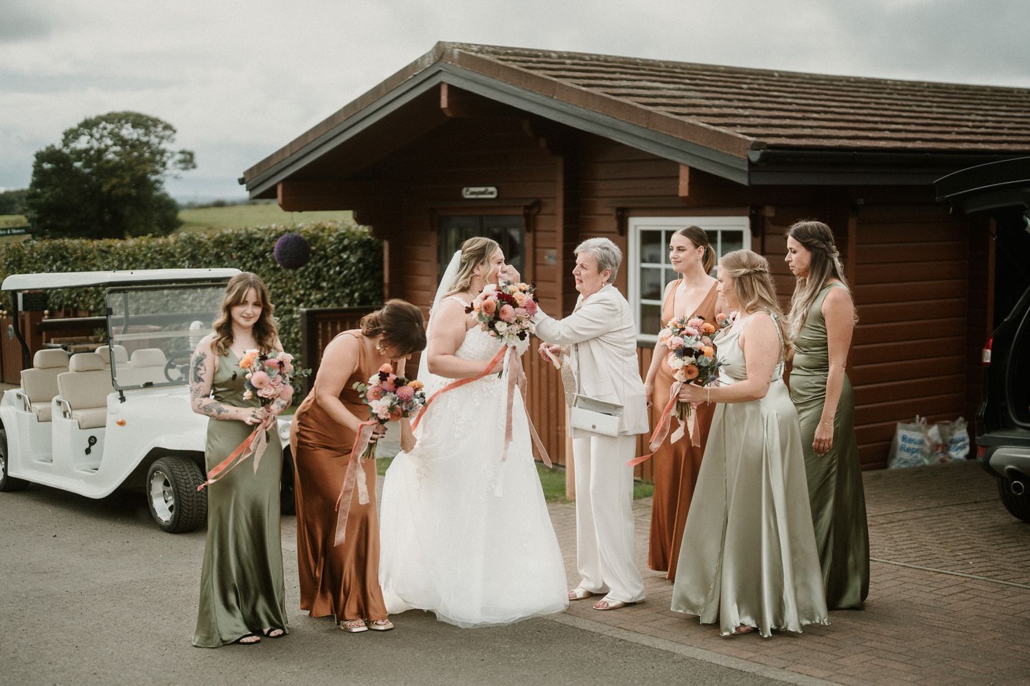 Wedding group in mix of sage green and rust colored dresses poses outside venue.