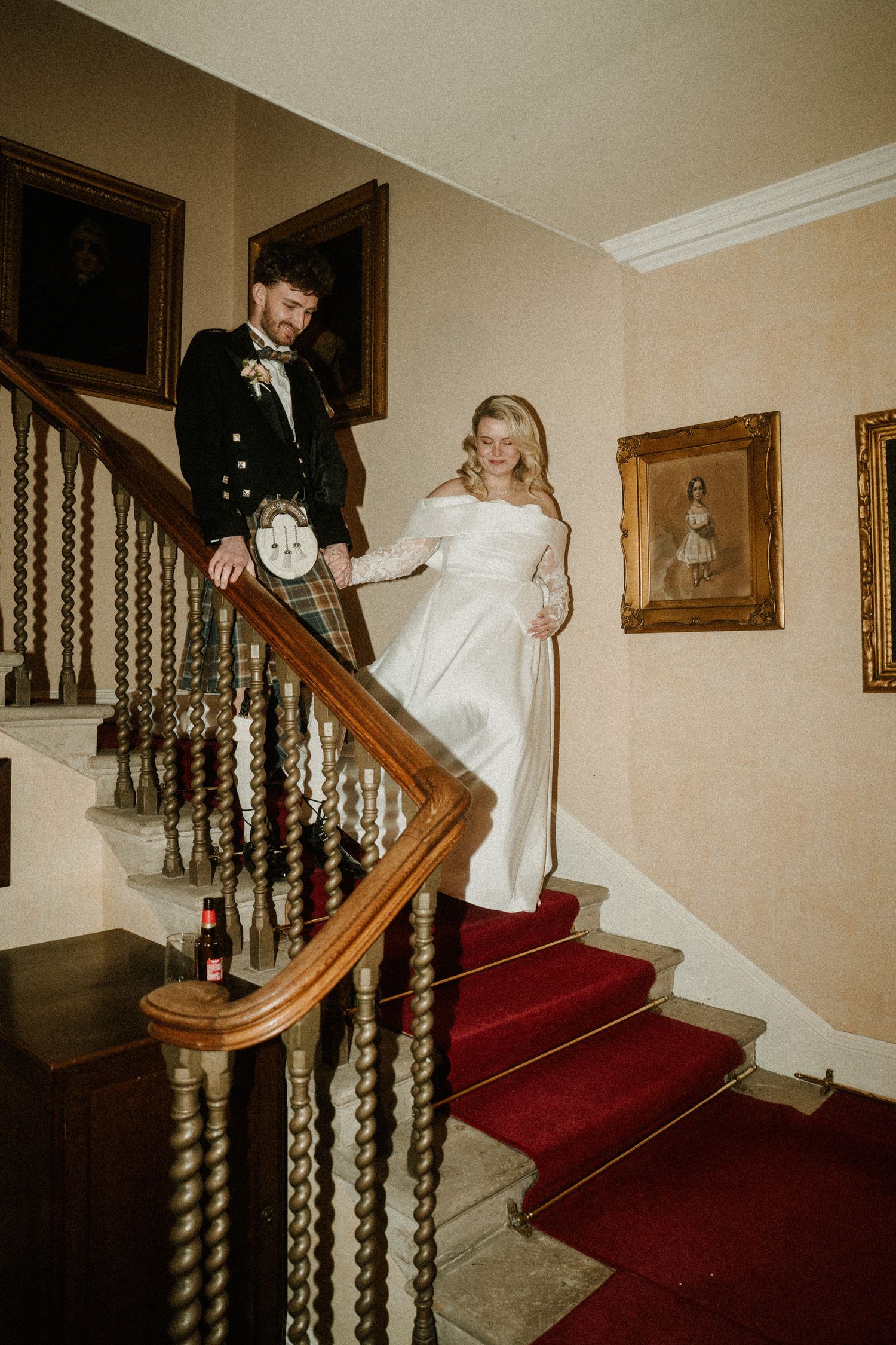 Wedding couple descending elegant staircase with traditional Scottish attire and white gown.