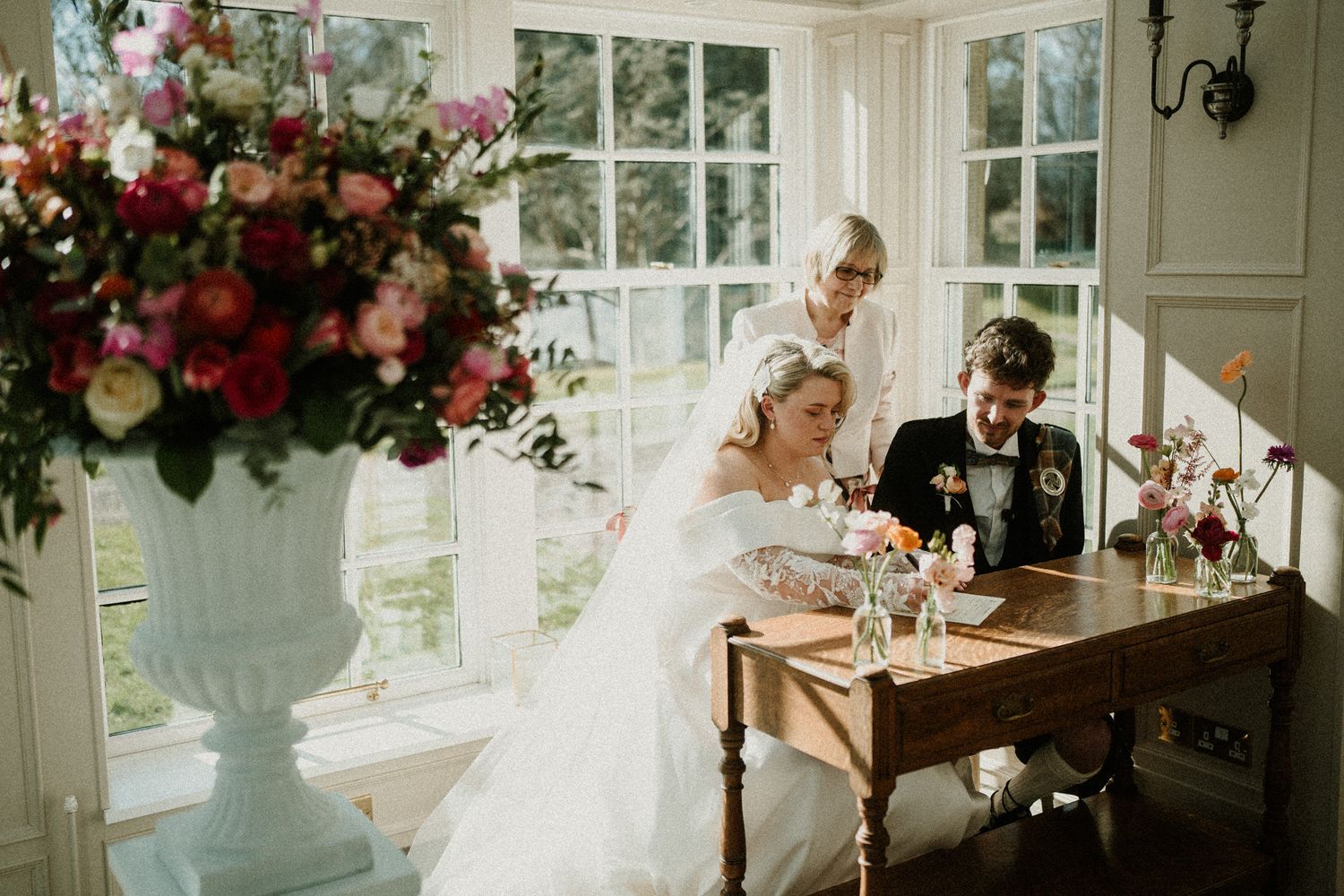 Couple signs marriage documents at an elegant wooden table decorated with vibrant floral arrangements.
