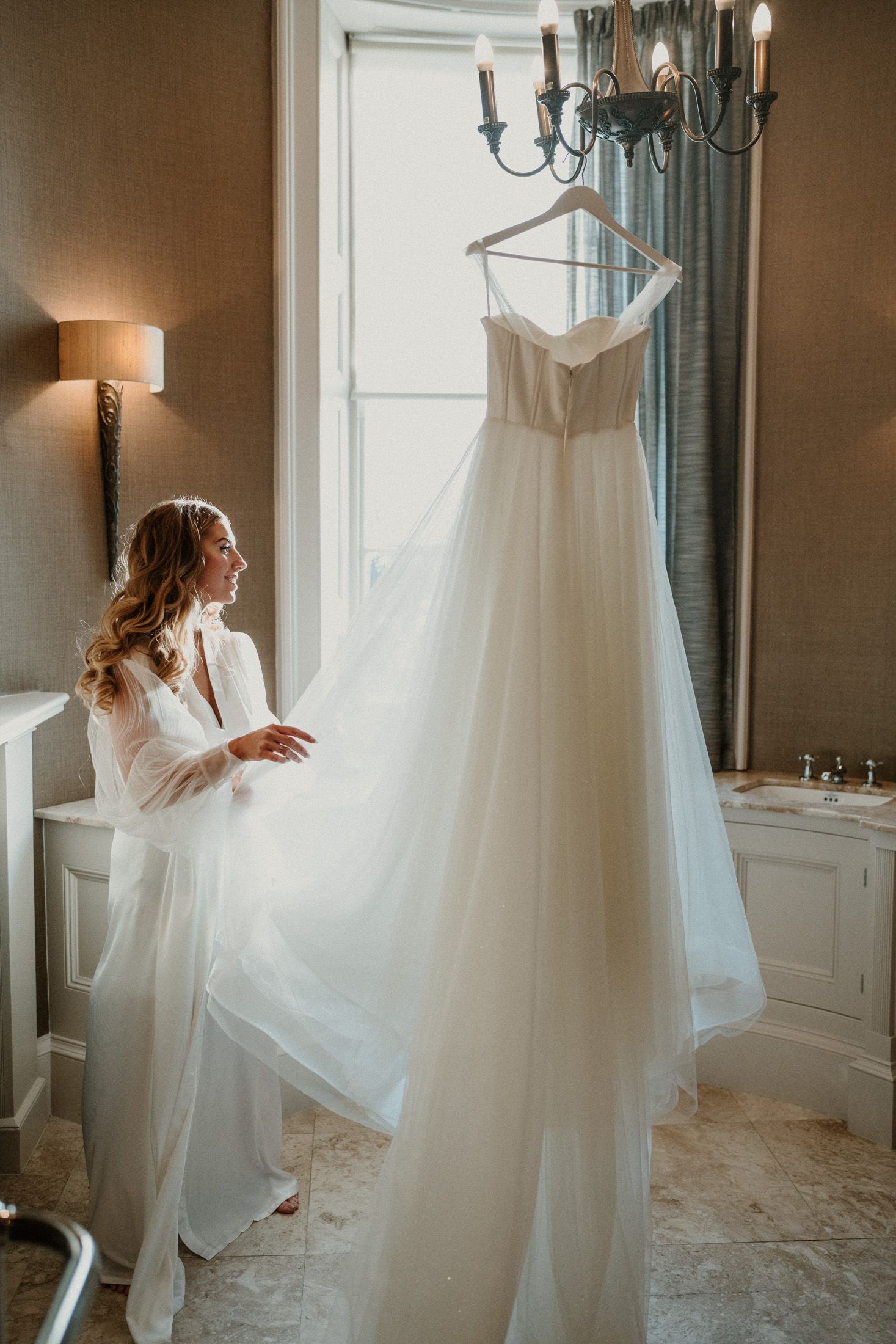 A white wedding dress hangs elegantly from a chandelier in a softly lit dressing room.