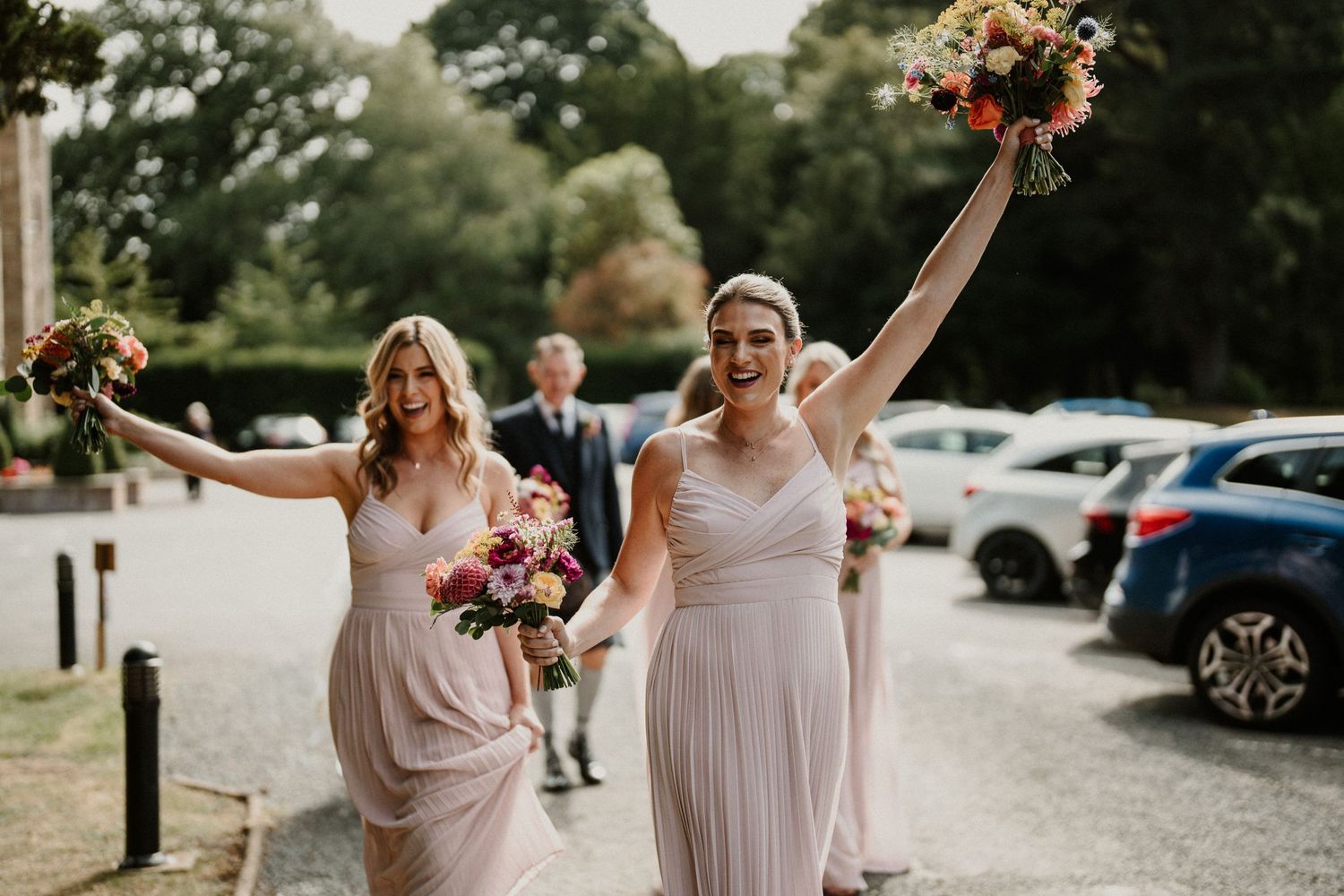 Wedding party celebrates joyfully outdoors in pale pink dresses holding floral bouquets.