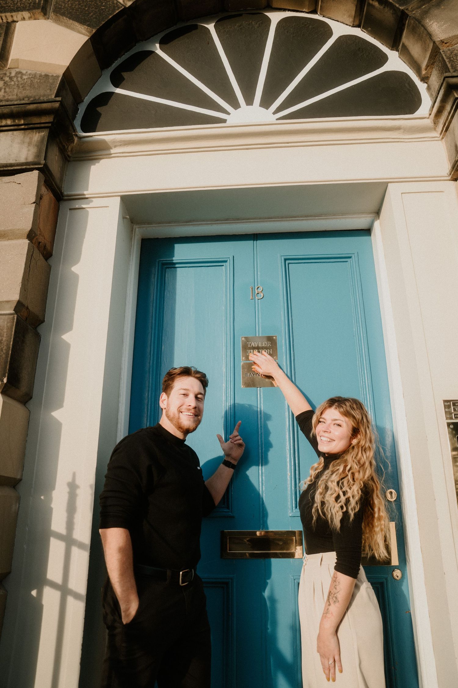 Couple smiling and knocking on a teal blue Georgian door with a decorative fanlight in Edinburgh.