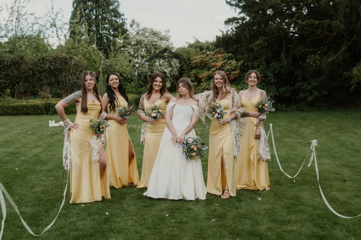 Bride in white gown with four bridesmaids in yellow dresses holding bouquets, posing on green lawn at outdoor wedding.