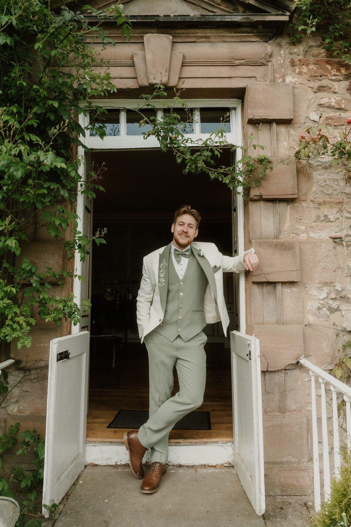 Groom in sage green suit and bow tie posing confidently in a stone doorway surrounded by lush greenery.