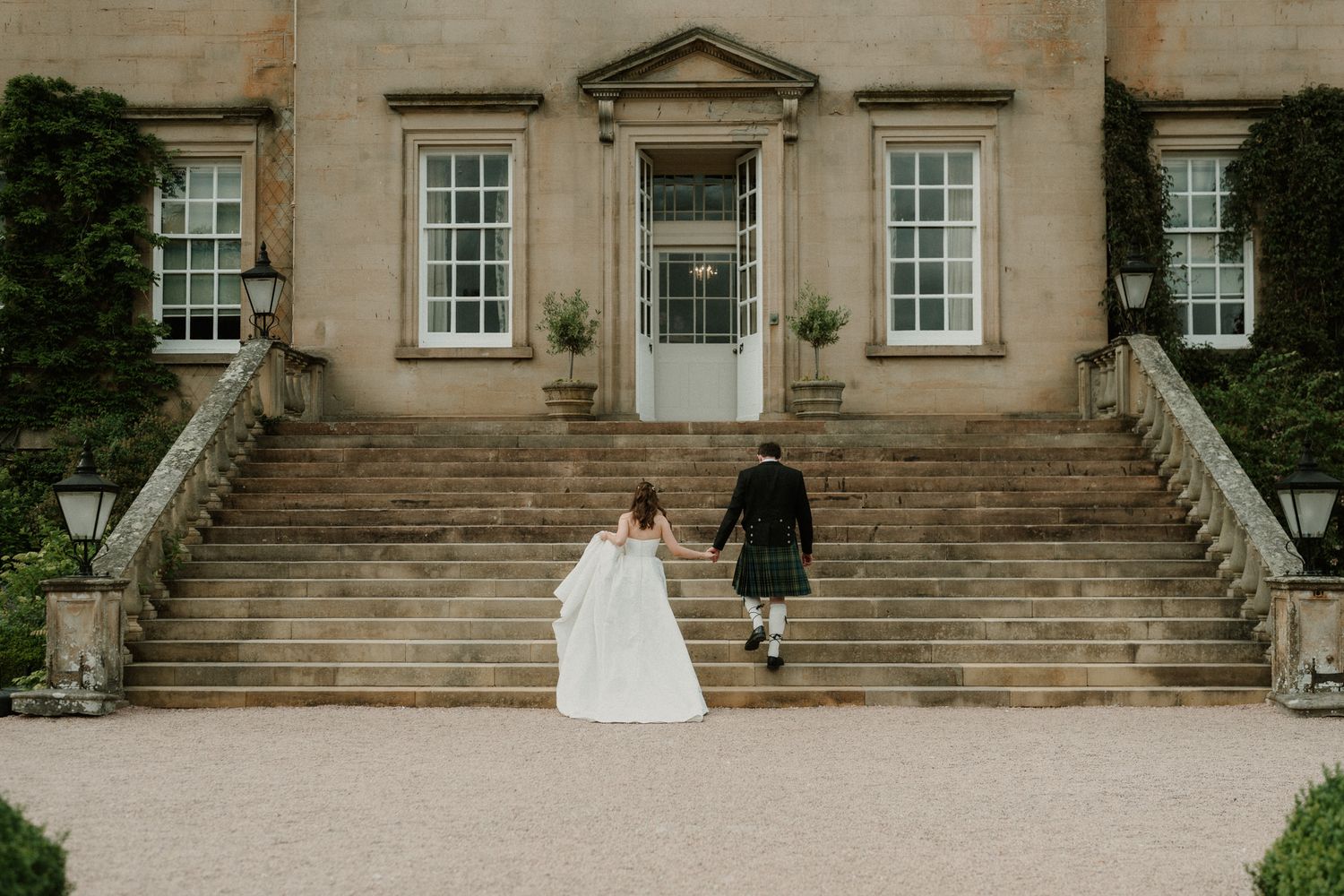 Bride and groom walking hand in hand up stone steps toward an elegant Scottish manor house wedding venue.