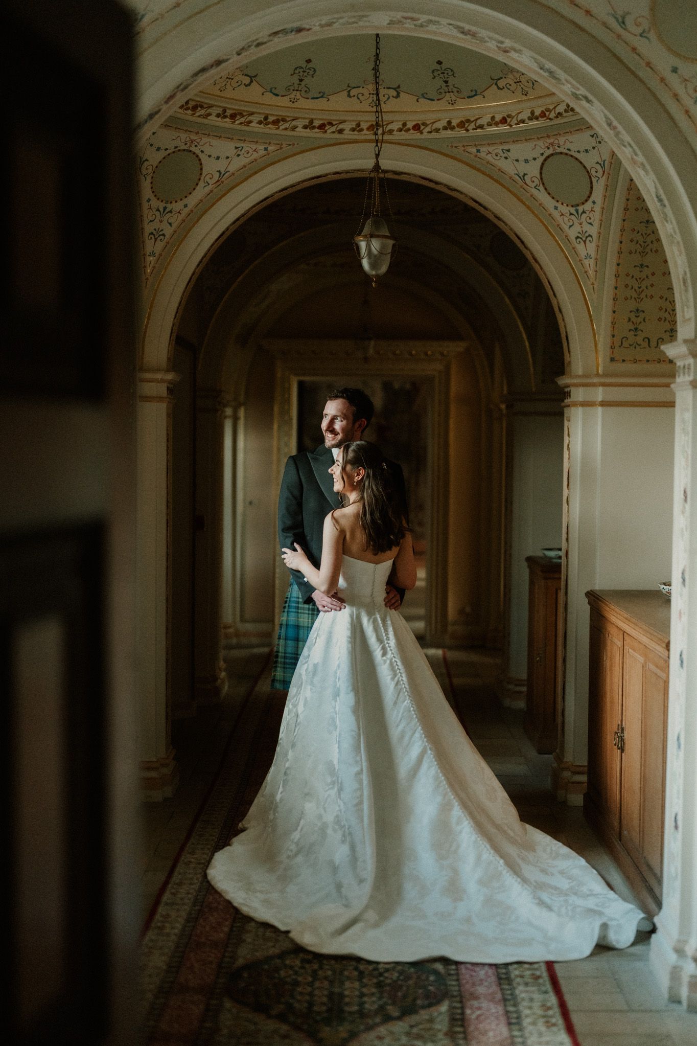 Bride and groom embrace in an ornate Scottish castle hallway, groom wearing green tartan kilt.