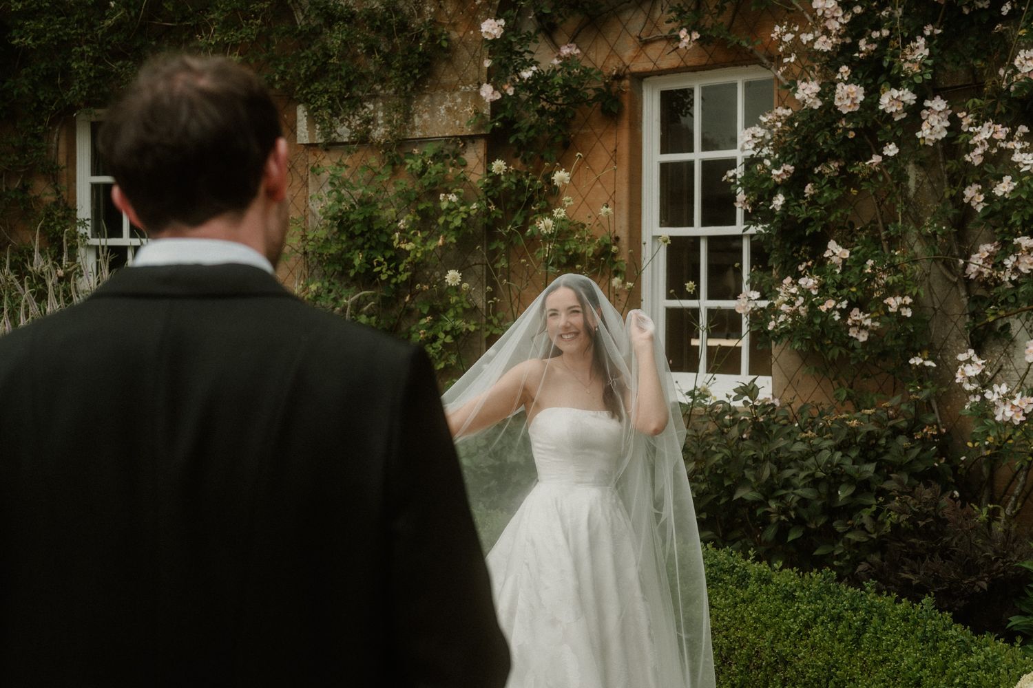 Bride in strapless gown and veil stands before a rose-covered stone manor, facing her groom during a romantic first look.