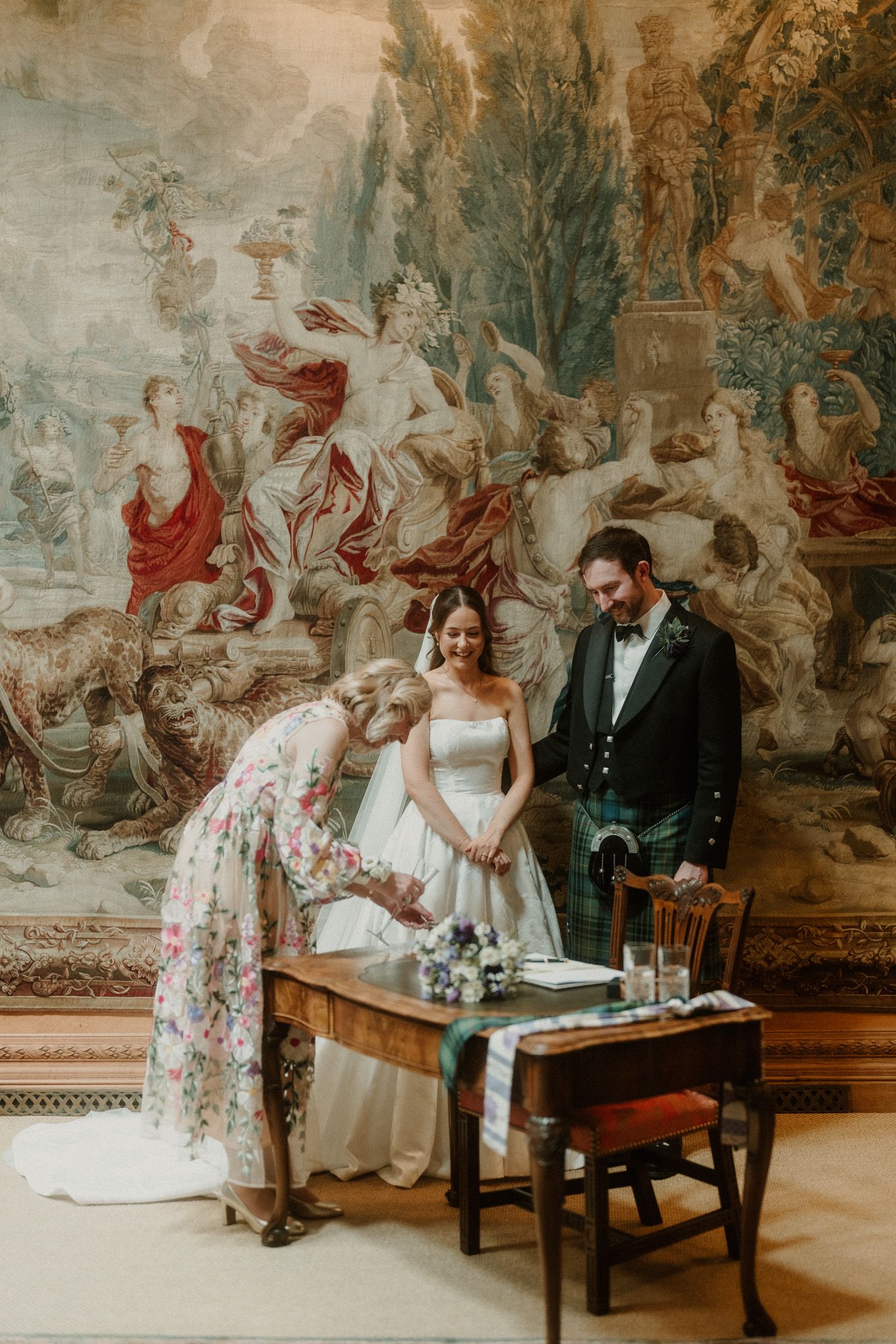 Bride signing wedding register at ornate wooden desk, groom in Scottish kilt, before grand tapestry backdrop.