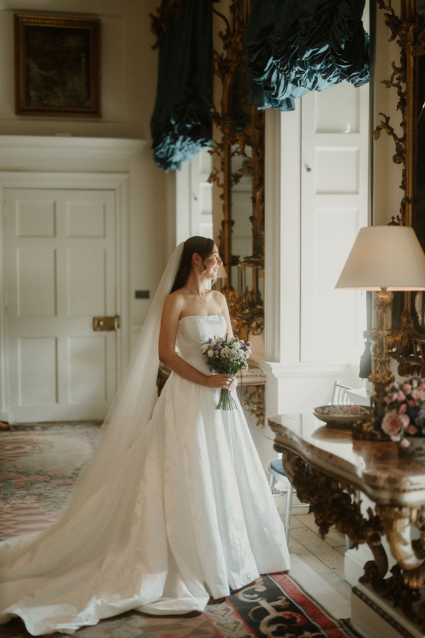 Bride in strapless white ball gown with veil holding bouquet stands by ornate gold mirror in elegant room.