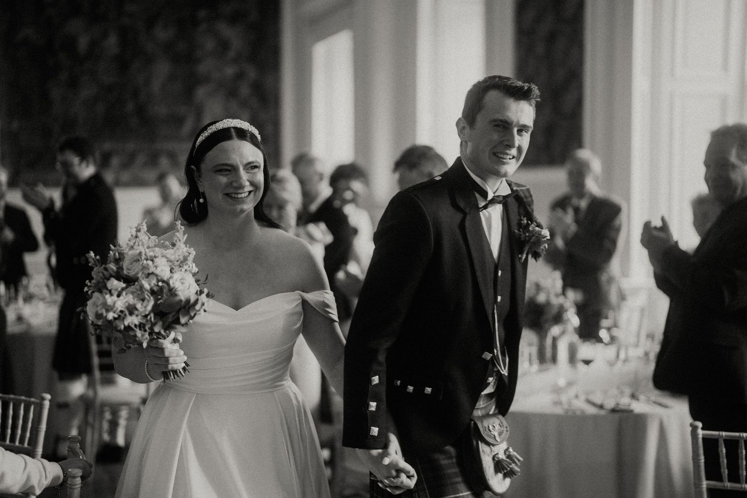 Wedding guests celebrate during an elegant reception in a grand ballroom with ornate chandeliers.