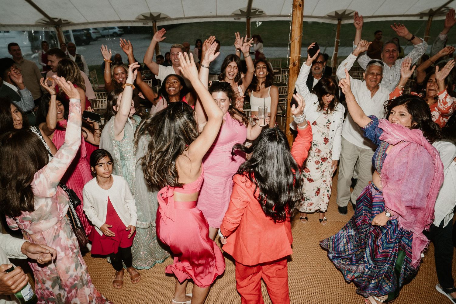 Energetic wedding reception guests dancing and celebrating with hands in the air under a white tent.