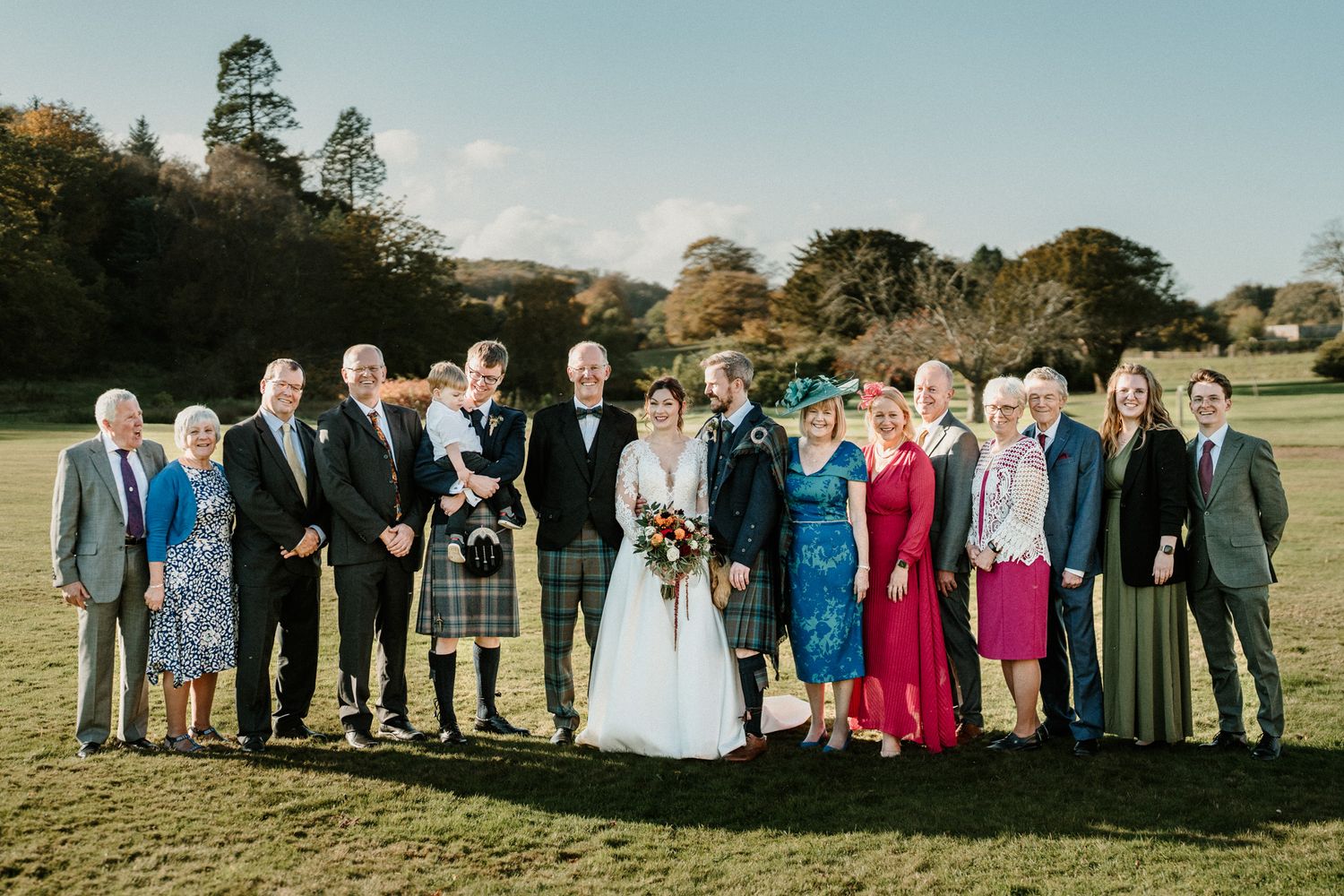 Wedding party and guests pose for formal group photo on sunny lawn with Scottish kilts.