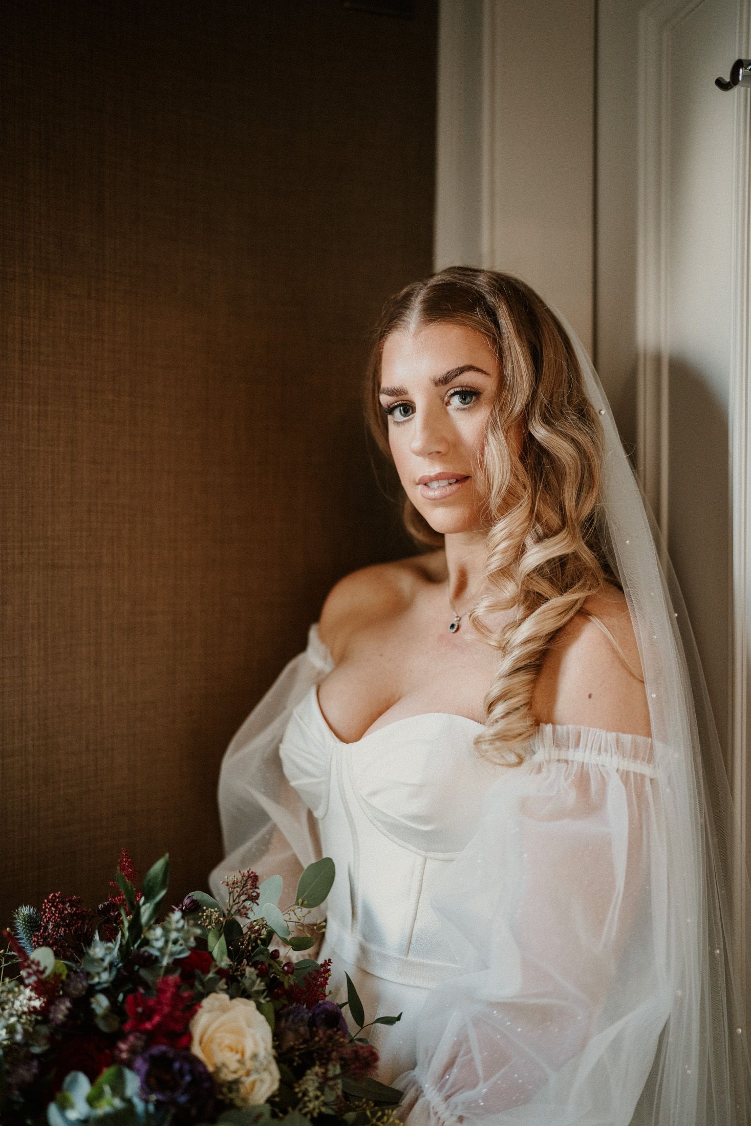 A bride holds a vibrant burgundy and white bouquet while posing against a textured wall.