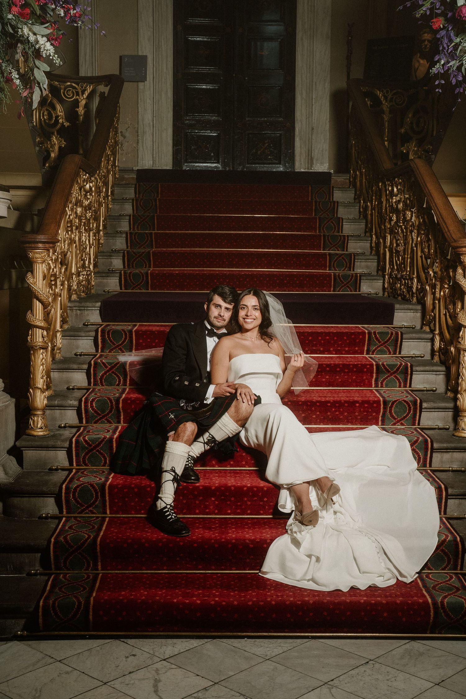 Couple sits on elegant red-carpeted stairs with ornate gold banisters.