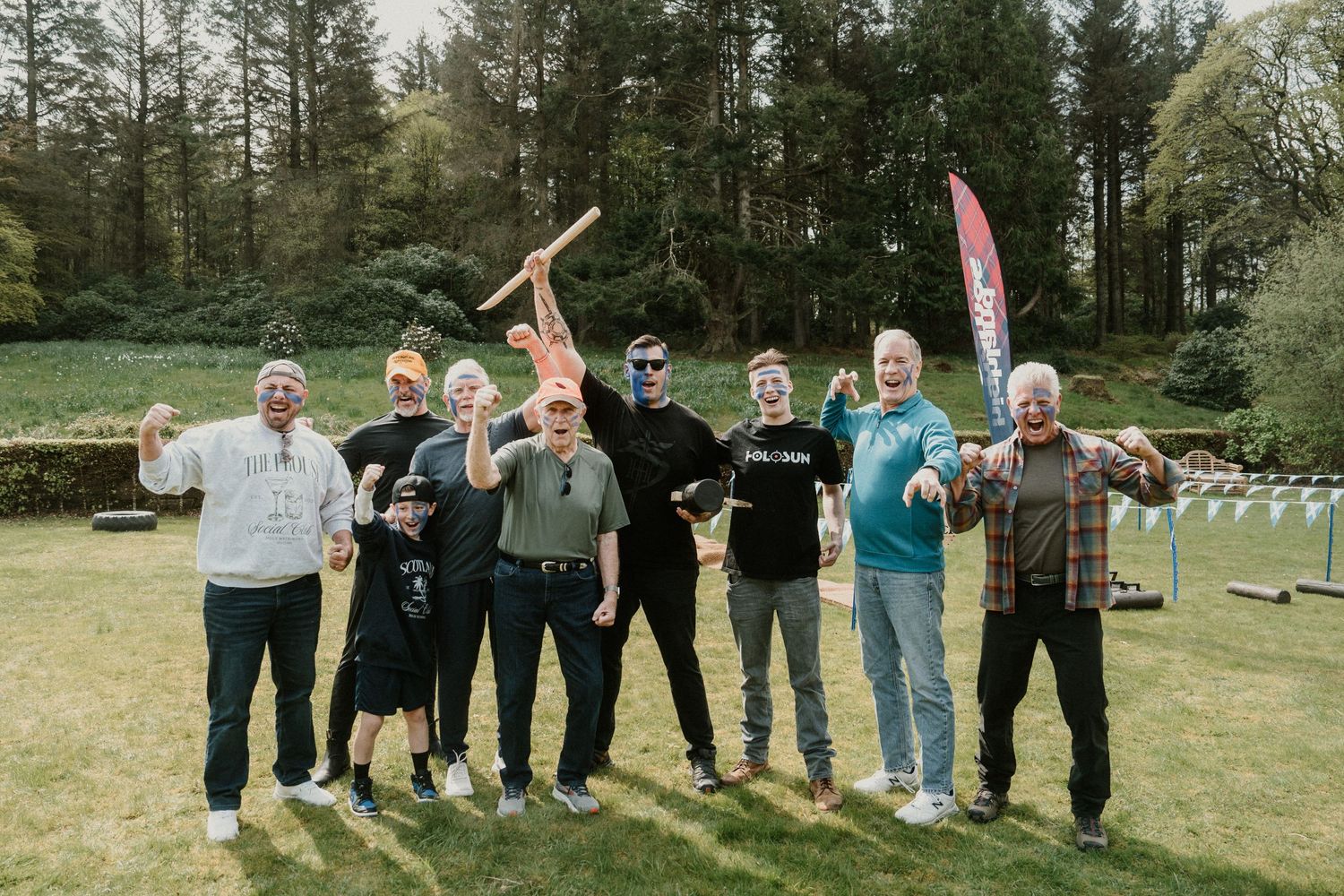 A group of people pose together holding axes on a grassy field with forest in background.