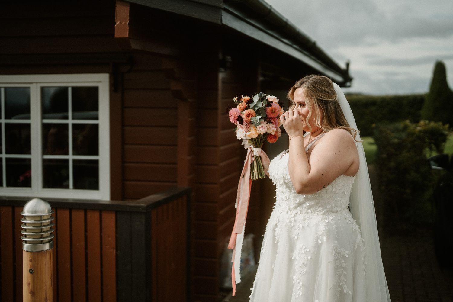 Wedding party members hold bouquets while standing in a line against wooden building exterior.
