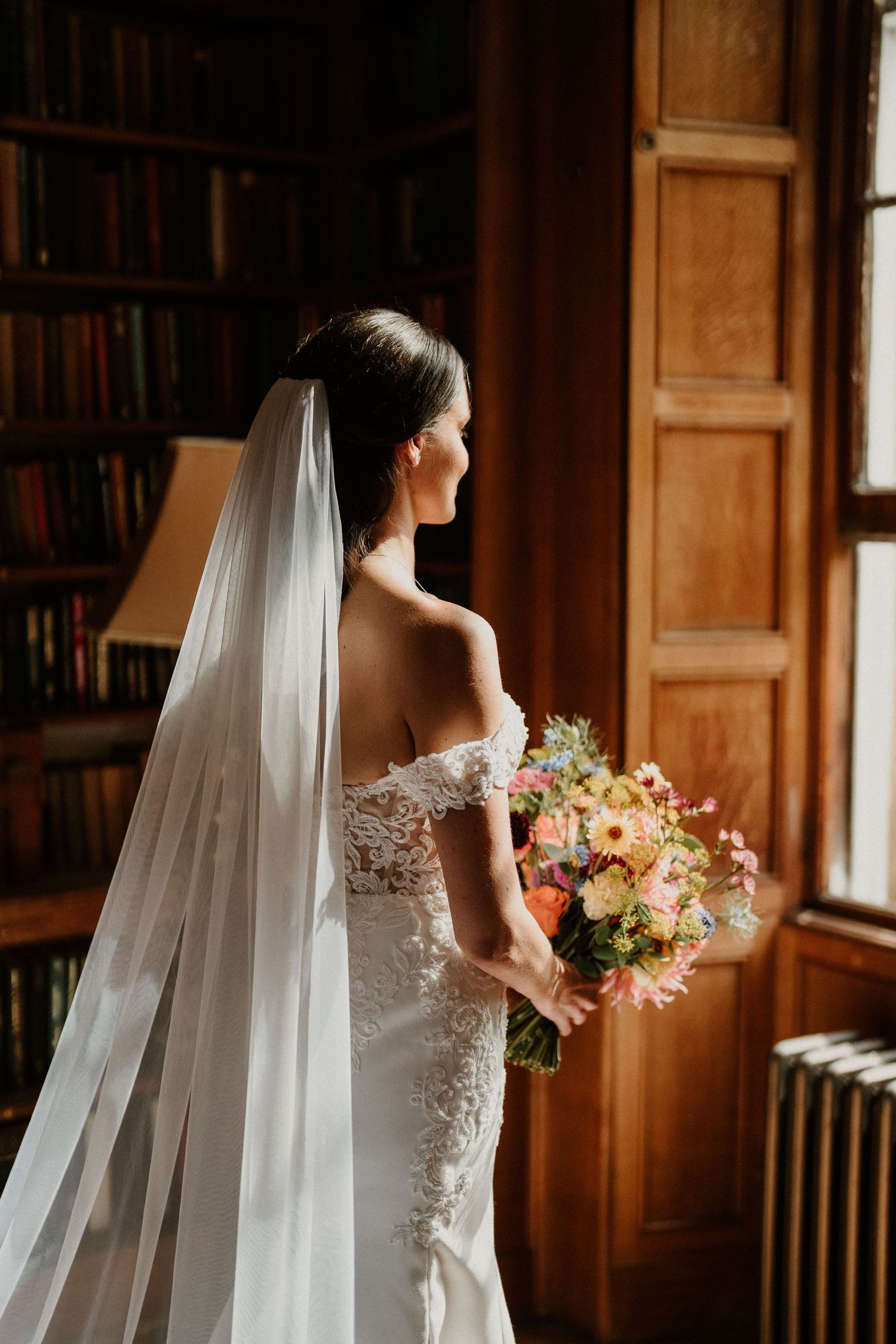 Bride holding colorful bouquet poses in elegant library setting with flowing veil.