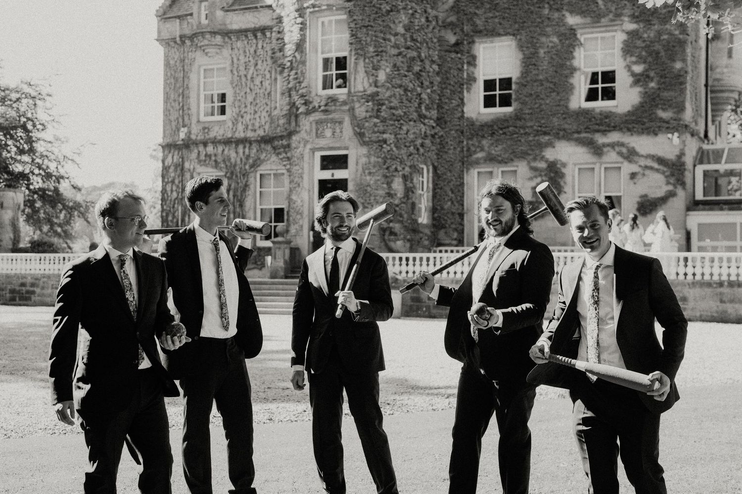 Groomsmen in black tuxedos pose playfully outside historic building in black and white photography.