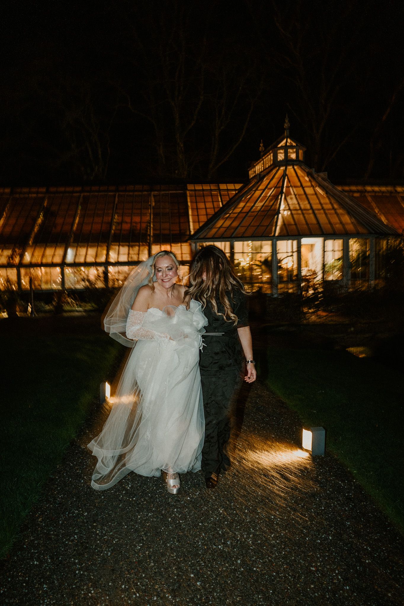 Wedding couple walks at night in front of illuminated greenhouse buildings creating a romantic atmosphere.